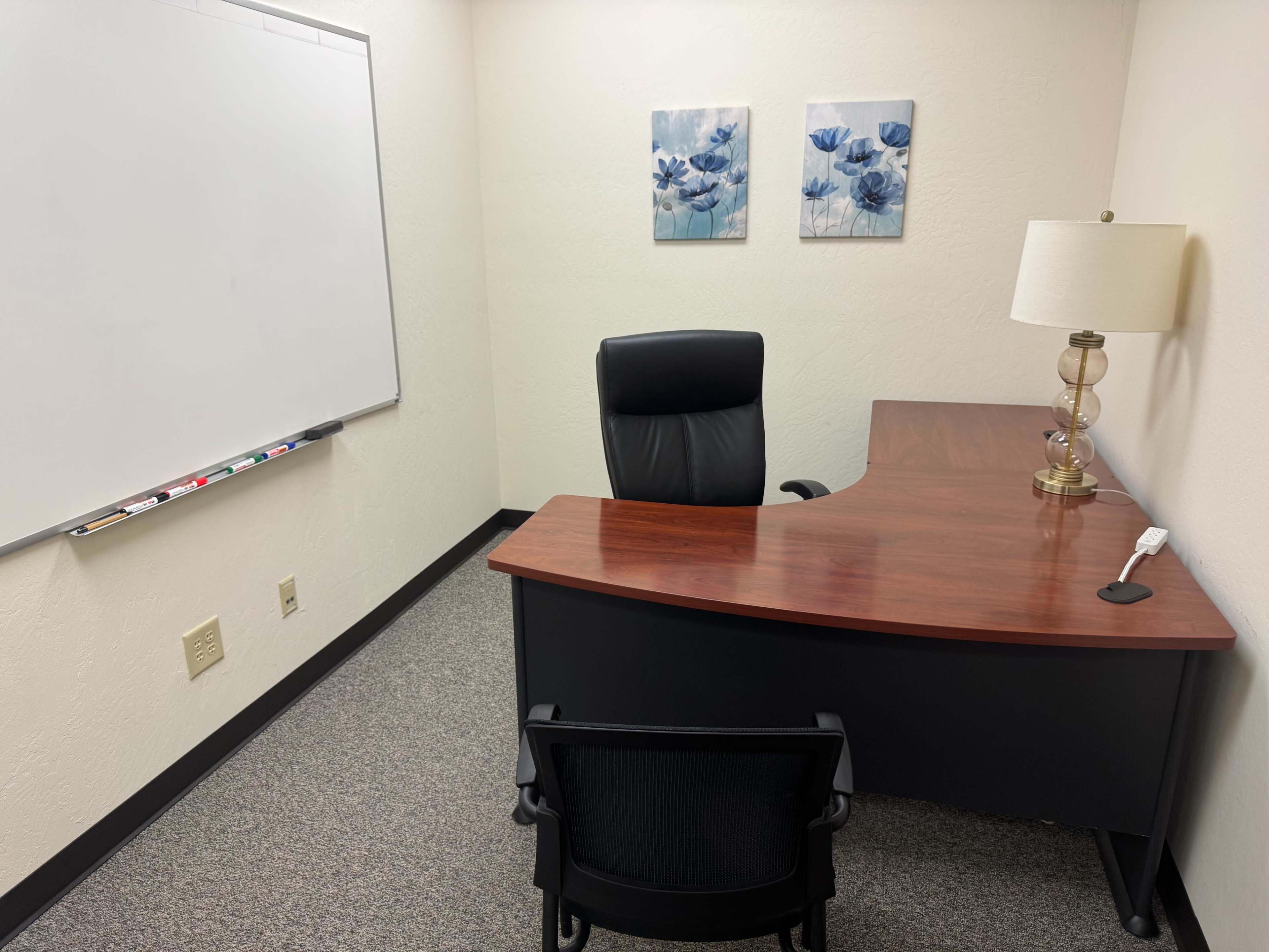 The image shows a small office with a wooden desk, a black office chair, a lamp, and two floral pictures on the wall, along with a whiteboard.