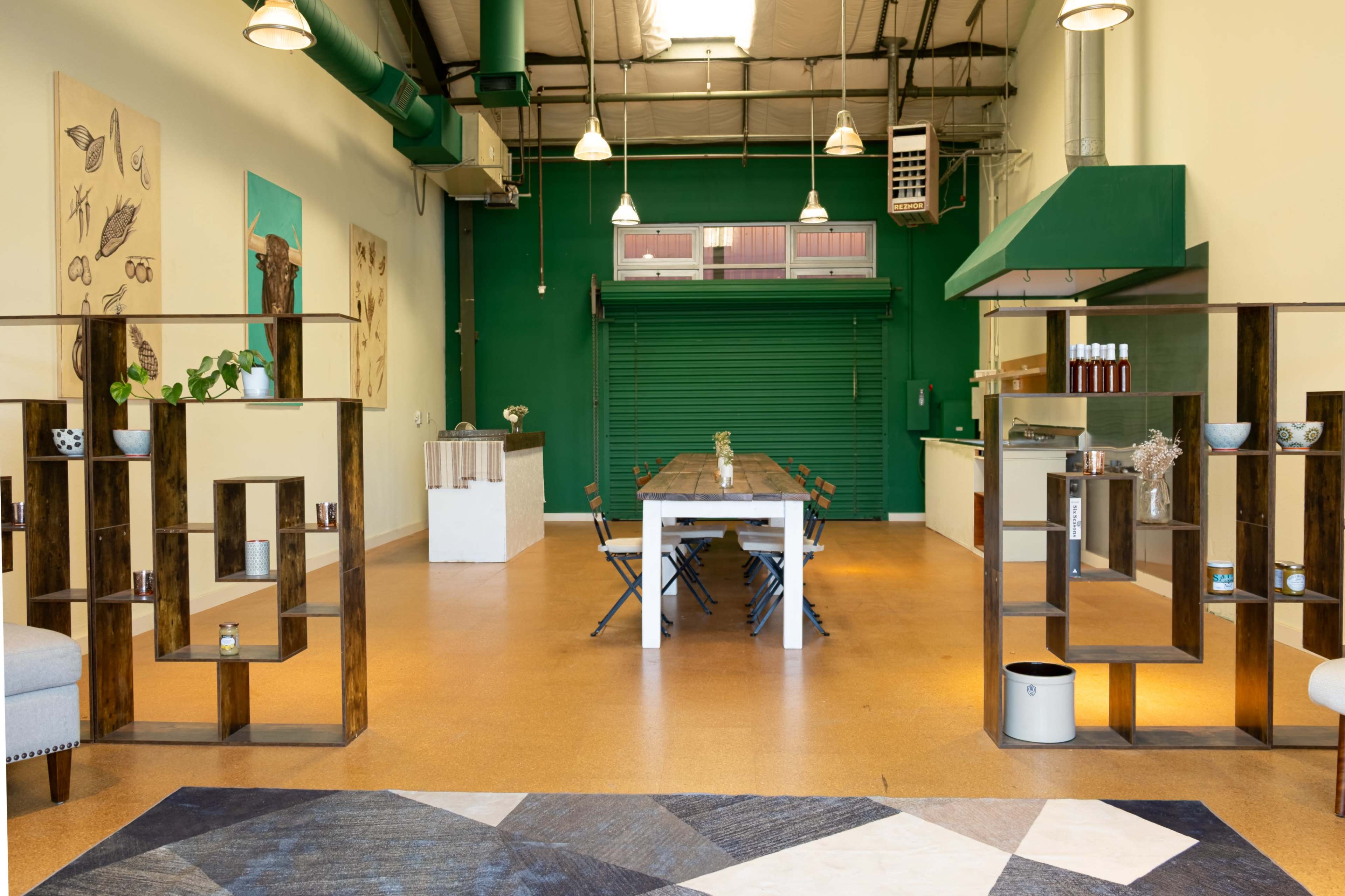 The image shows a spacious interior featuring a large white table surrounded by chairs, wooden display shelves, and green walls with a closed roll-up door in the background.