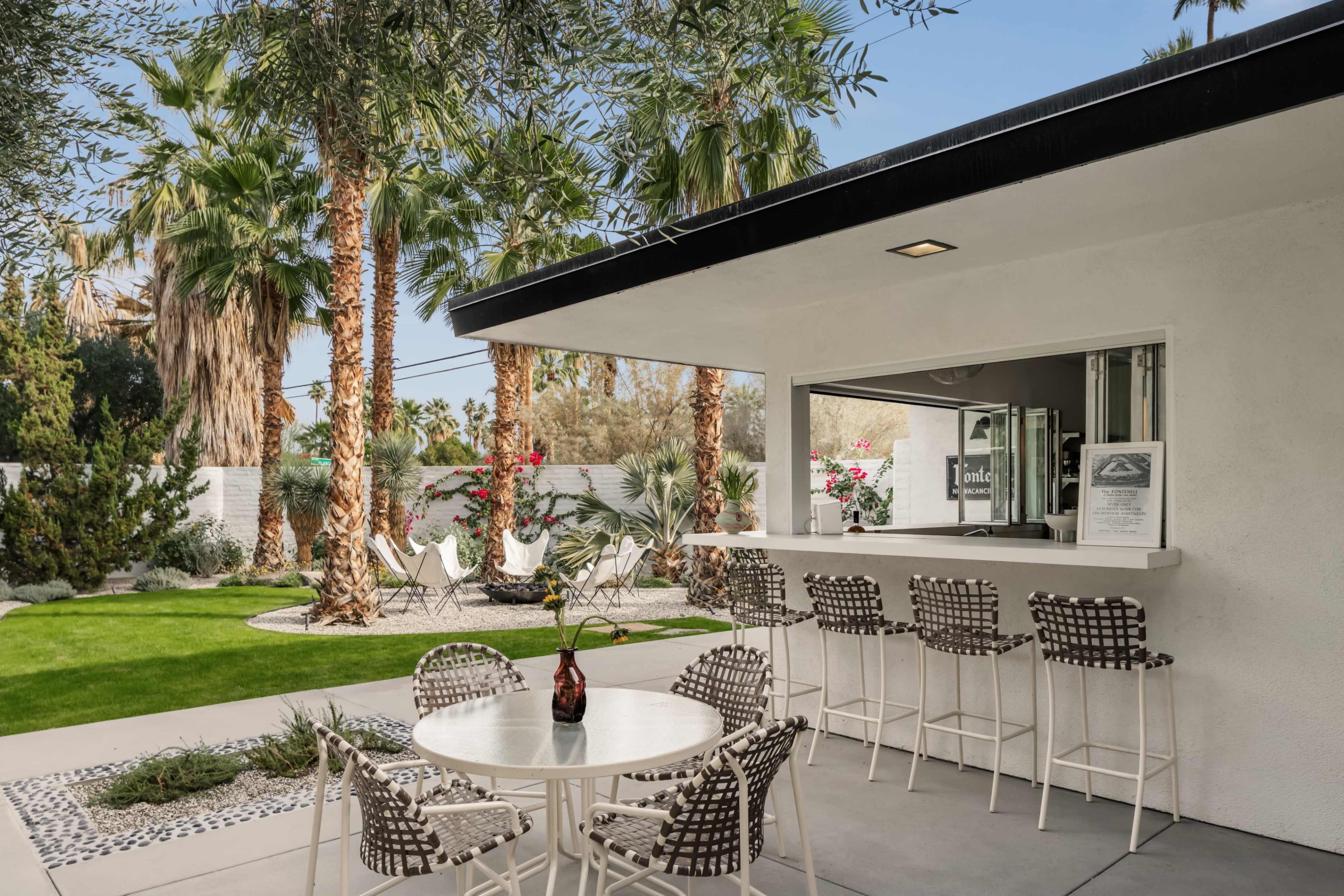 A modern outdoor seating area features a bar with high stools, a round table, and lush palm trees in the background.