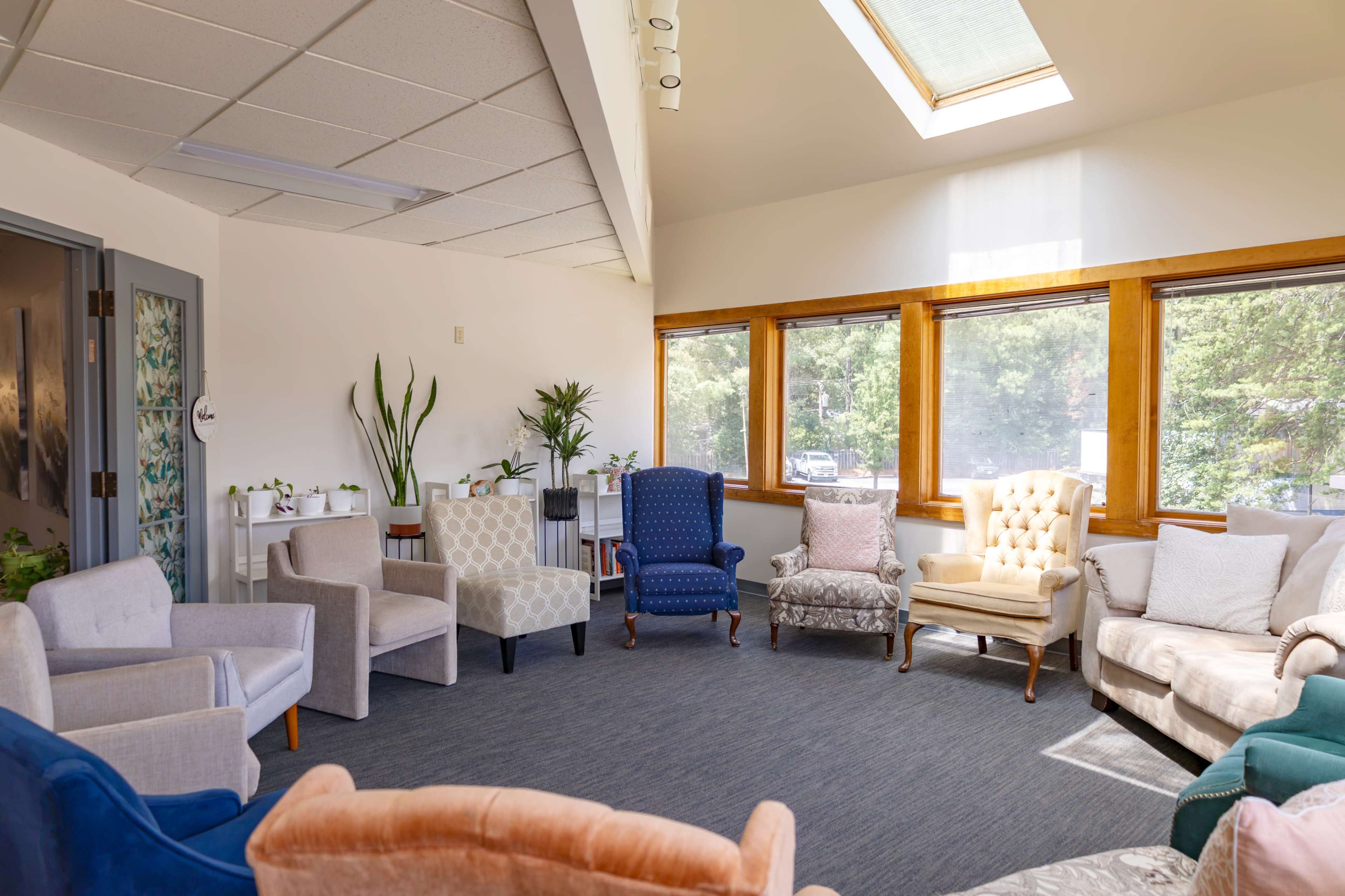 The image shows a brightly lit room with large windows, featuring a circle of various upholstered chairs and a few indoor plants.