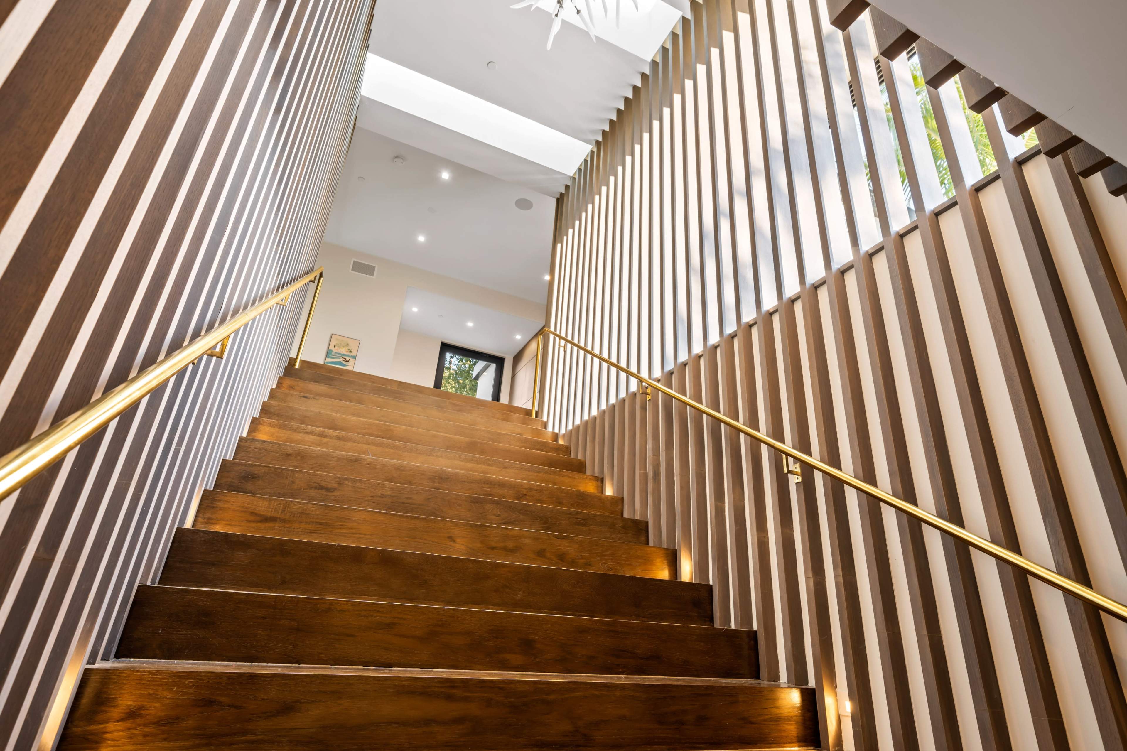 A staircase with wooden steps and a gold railing leads upward through slatted wooden walls to a well-lit landing.