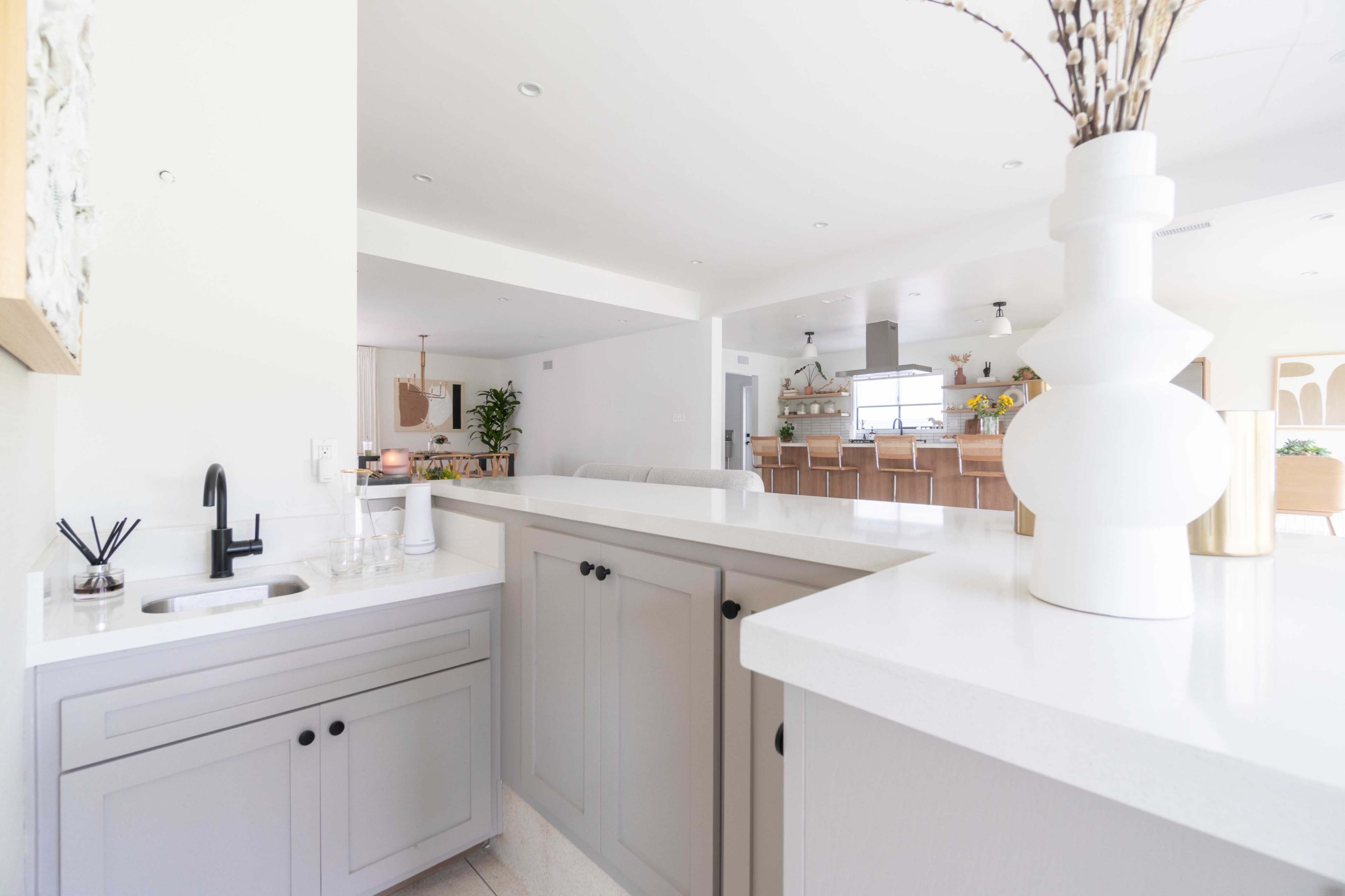 A modern kitchen features a gray cabinetry with a white countertop, displaying a sink and decorative vase, leading into an open dining area.