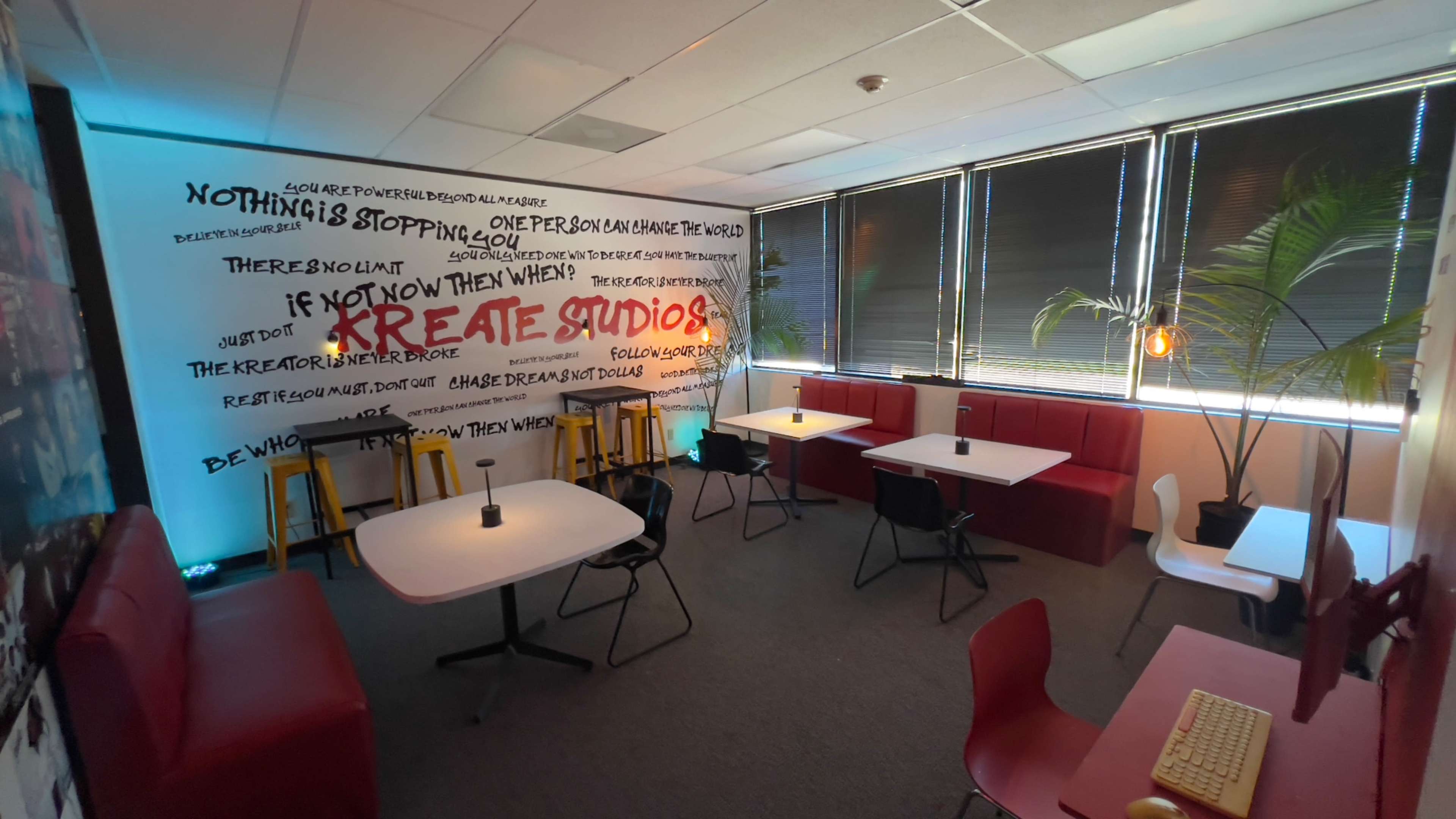 A creative workspace features white tables, chairs, and a large wall mural with motivational text, surrounded by bright lighting and greenery.
