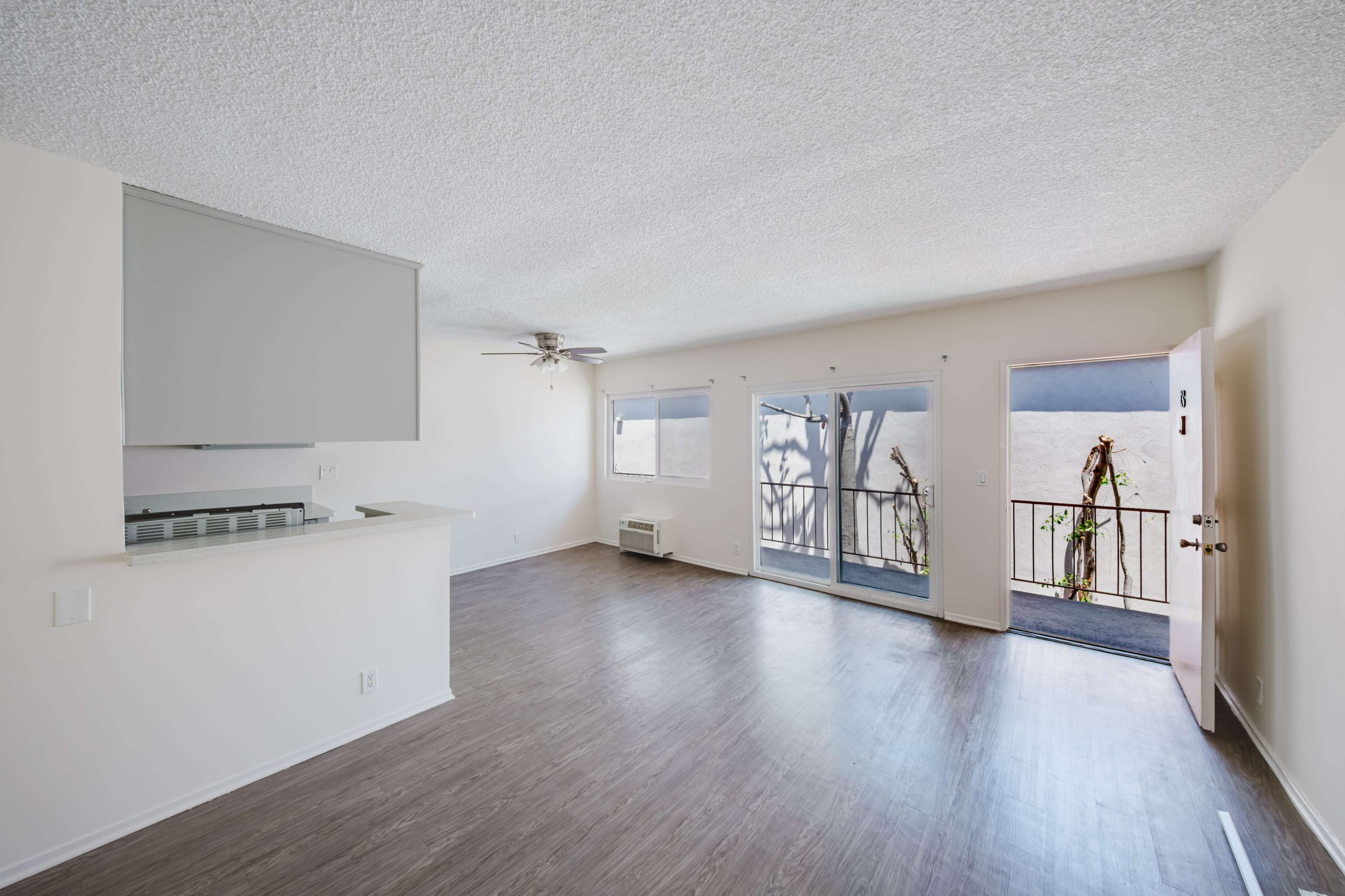 The image shows a spacious, empty living area with a ceiling fan, a sliding glass door leading to a balcony, and an open kitchen layout.