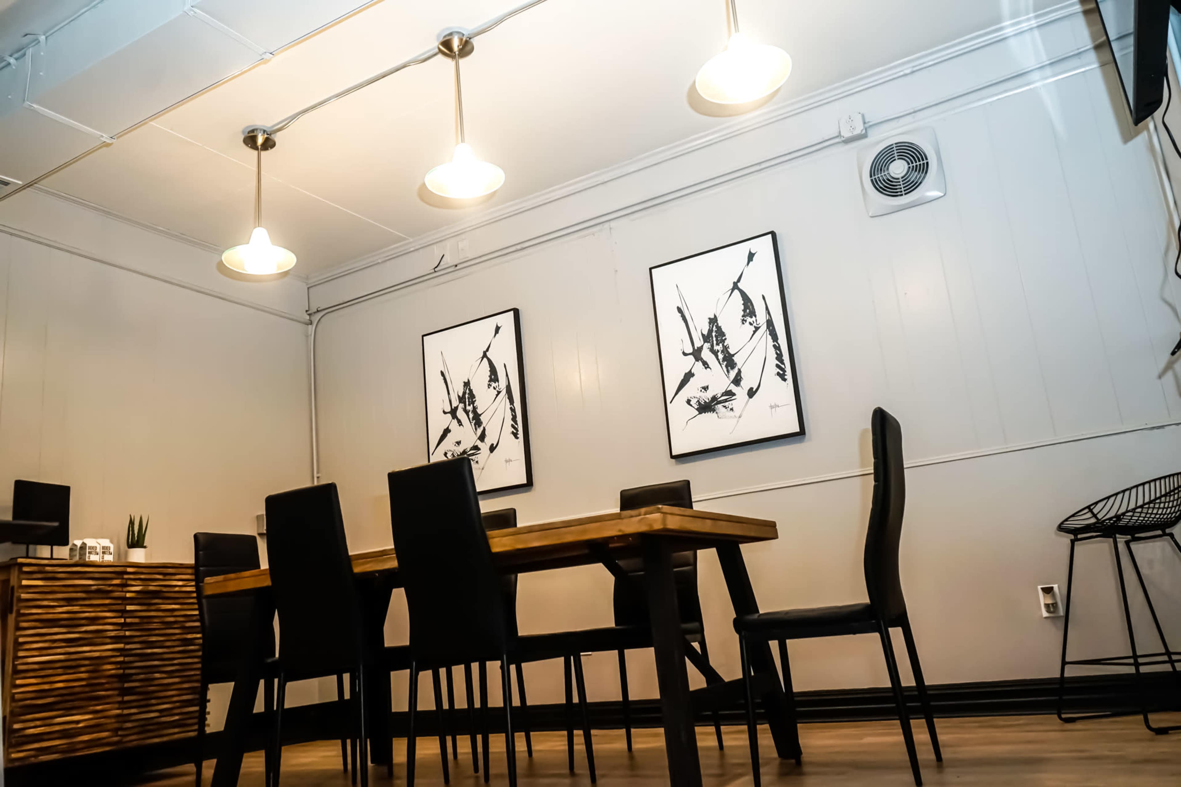 A minimalist meeting room features a rectangular wooden table surrounded by black chairs, with two framed art pieces on the wall and pendant lights above.