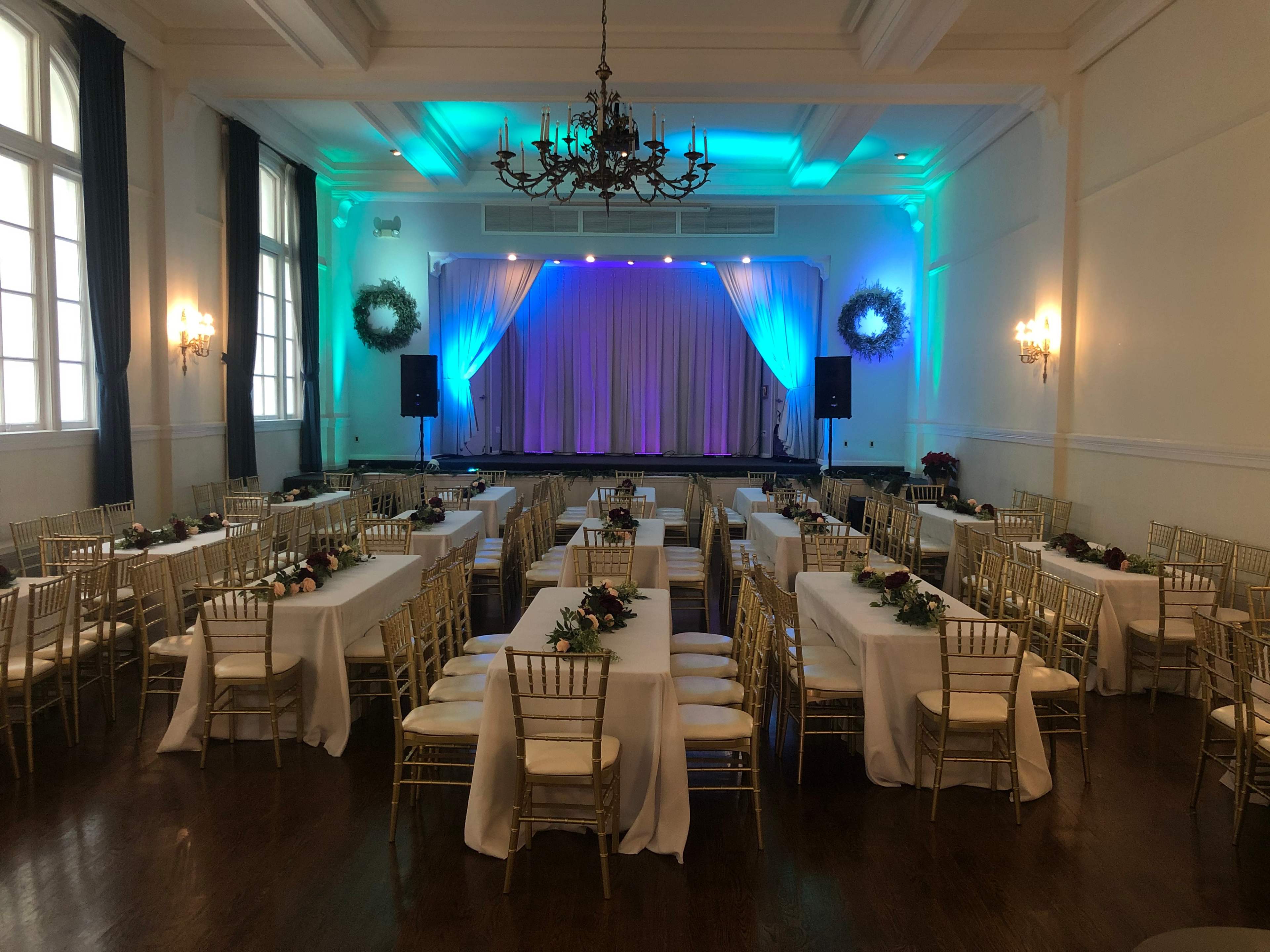 A banquet hall is set up with gold chairs and tables covered in white linens, featuring floral centerpieces, with a stage at the front adorned with draped fabric and lit in blue and purple tones.
