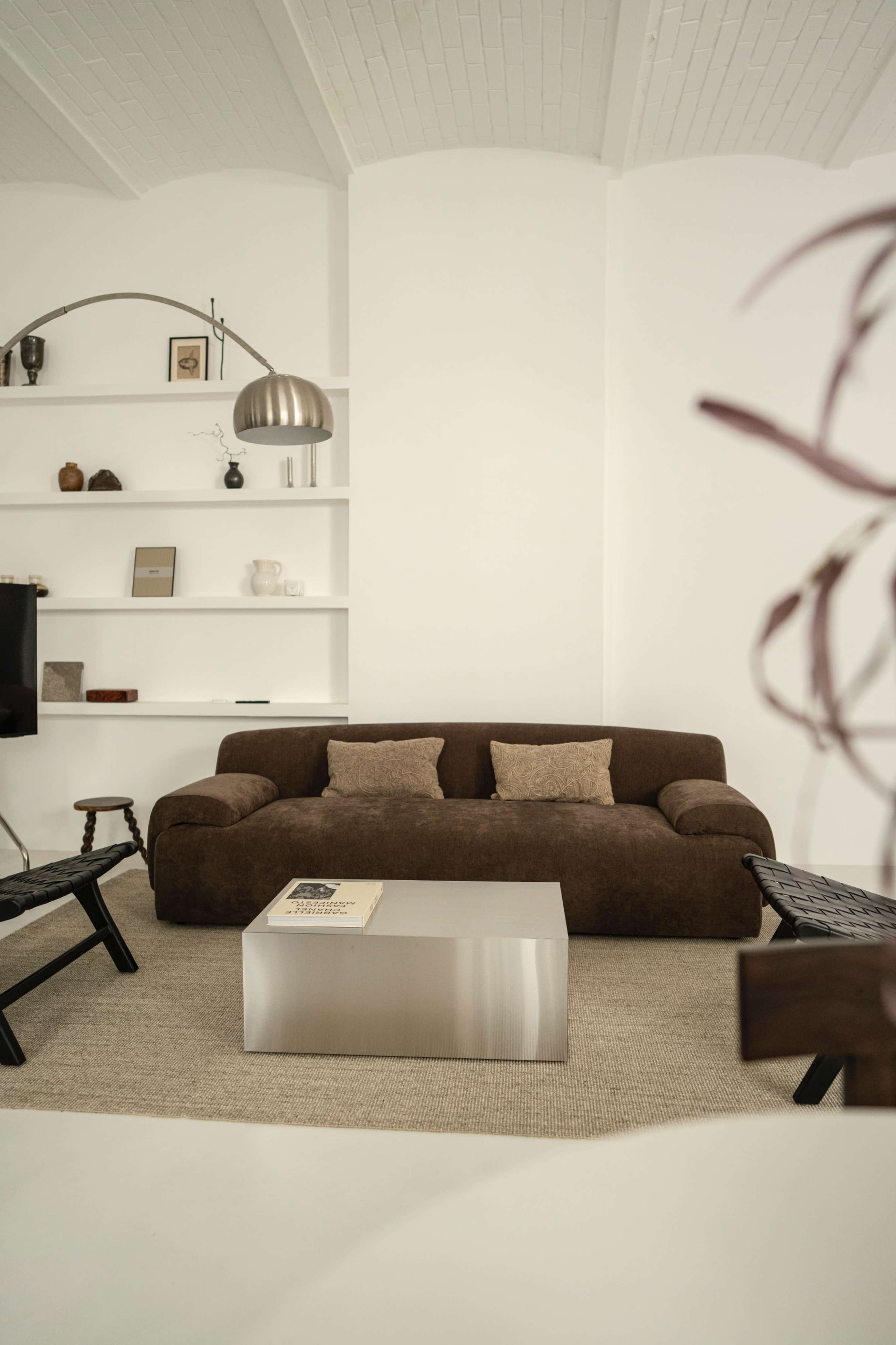 The image shows a minimalist living room featuring a brown sofa, a metallic coffee table, and a bookshelf adorned with various decorative objects.