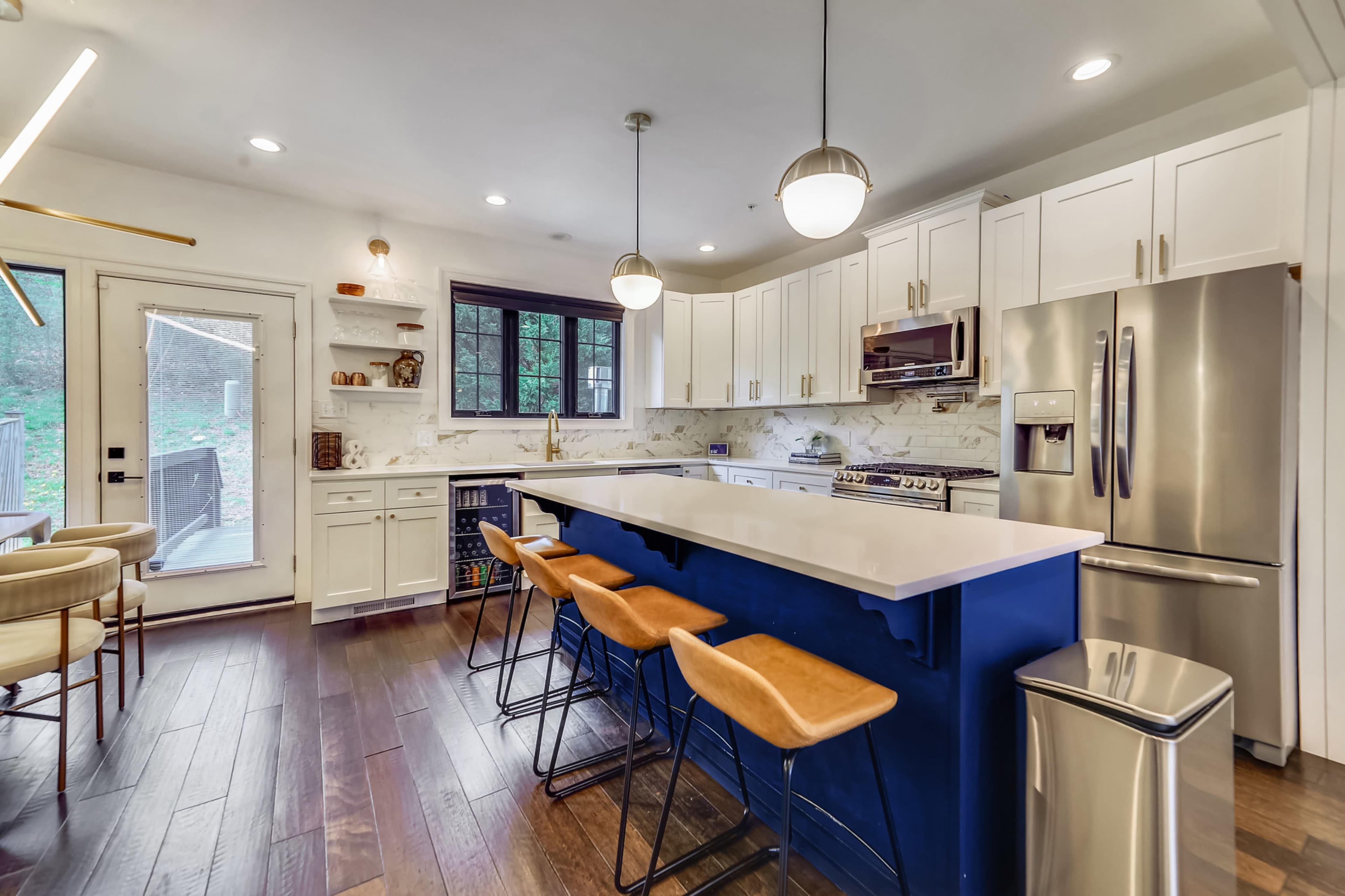 A modern kitchen featuring a blue island with bar stools, white cabinetry, stainless steel appliances, and large windows letting in natural light.