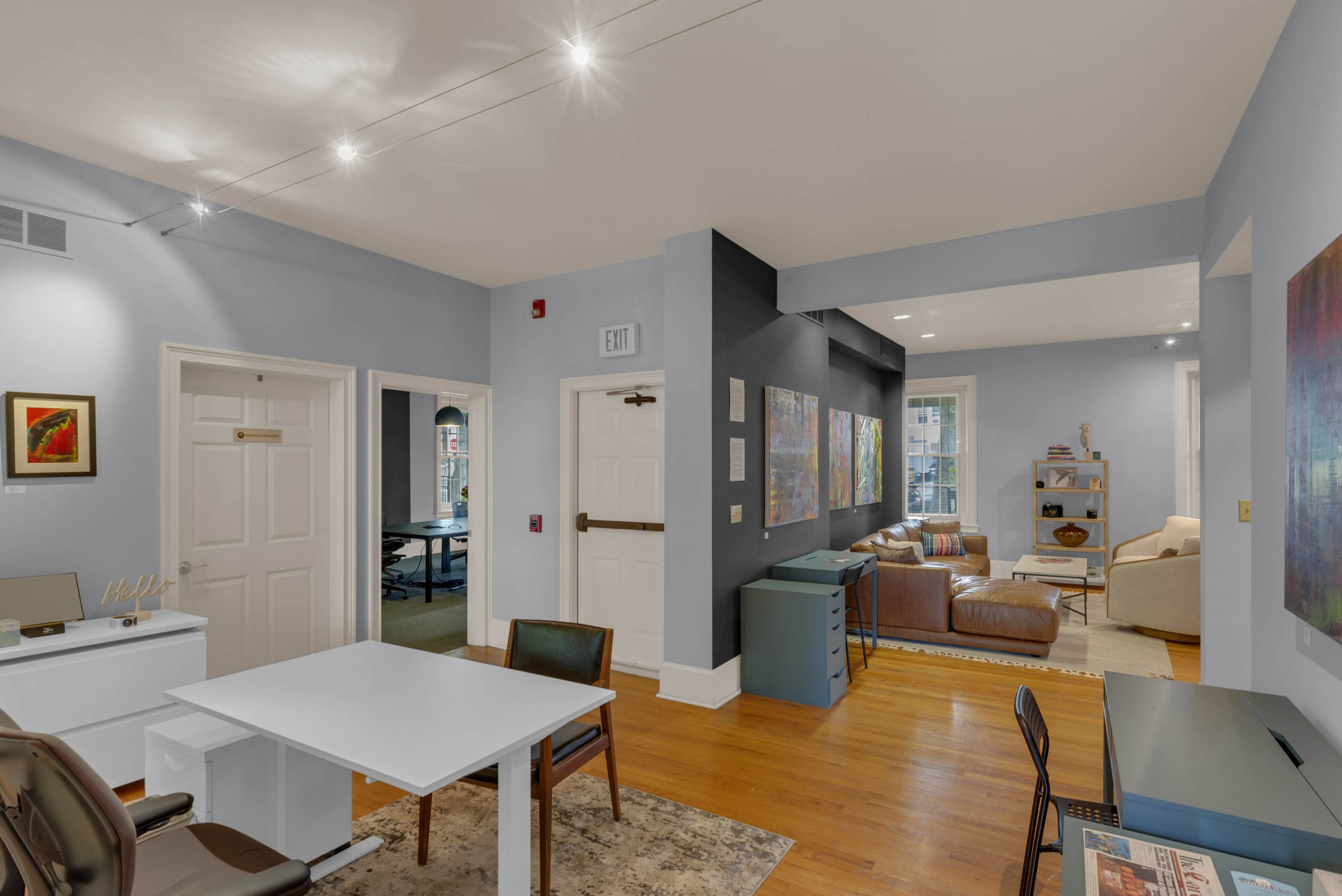 The image shows an interior space featuring a modern office area with a white desk and chair, leading into a cozy living area furnished with a brown sofa and additional seating.
