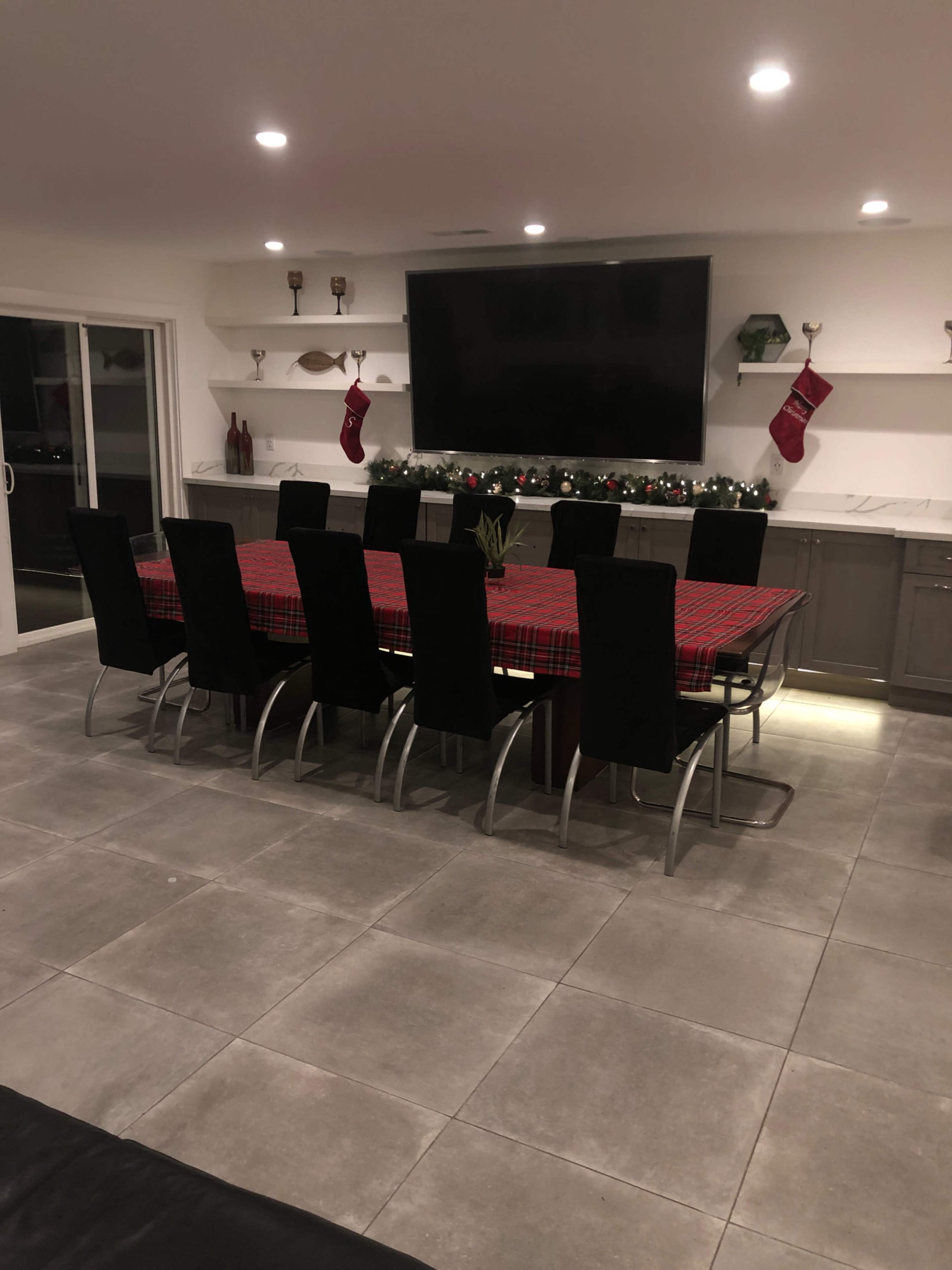 A long dining table covered with a red plaid tablecloth is set in a modern kitchen with seating for ten, and stockings are hung on the wall above a decorative mantel.