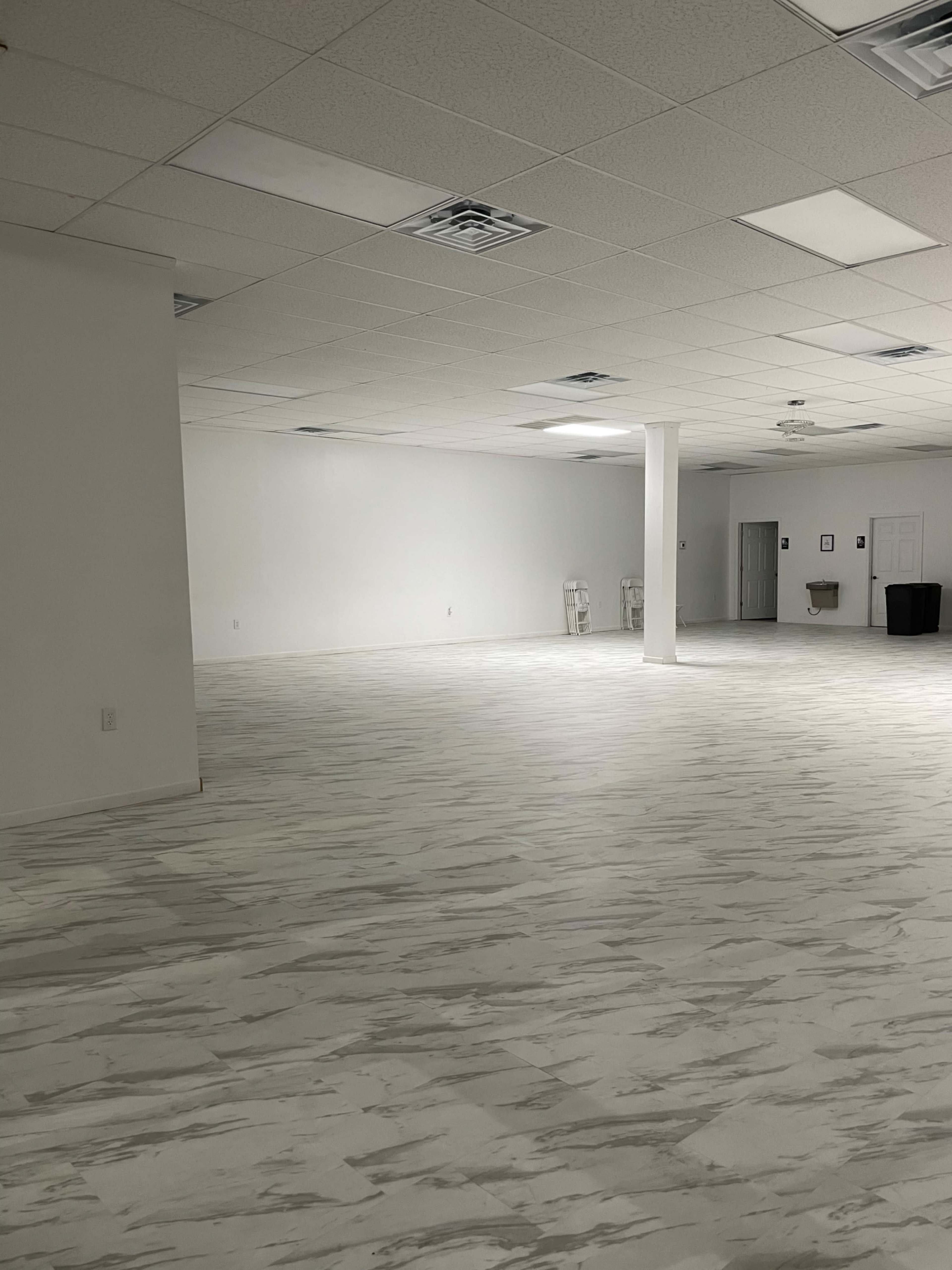 A spacious, empty room with white walls and a tiled floor, featuring minimal furniture and overhead lighting.