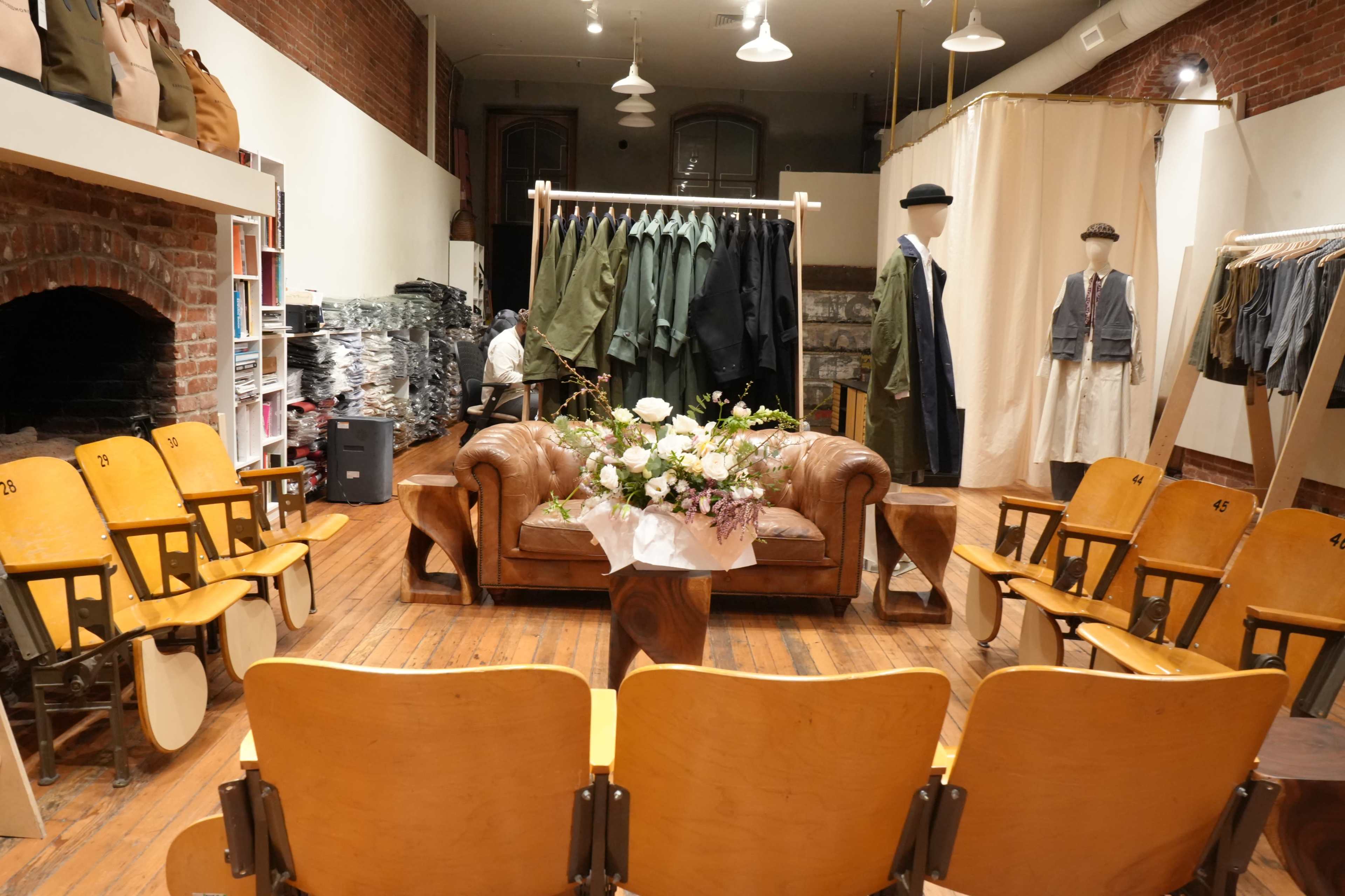 The image shows an interior of a clothing store featuring a brown leather sofa, a round coffee table with a flower arrangement, and rows of wooden chairs facing a clothing display on a rack.
