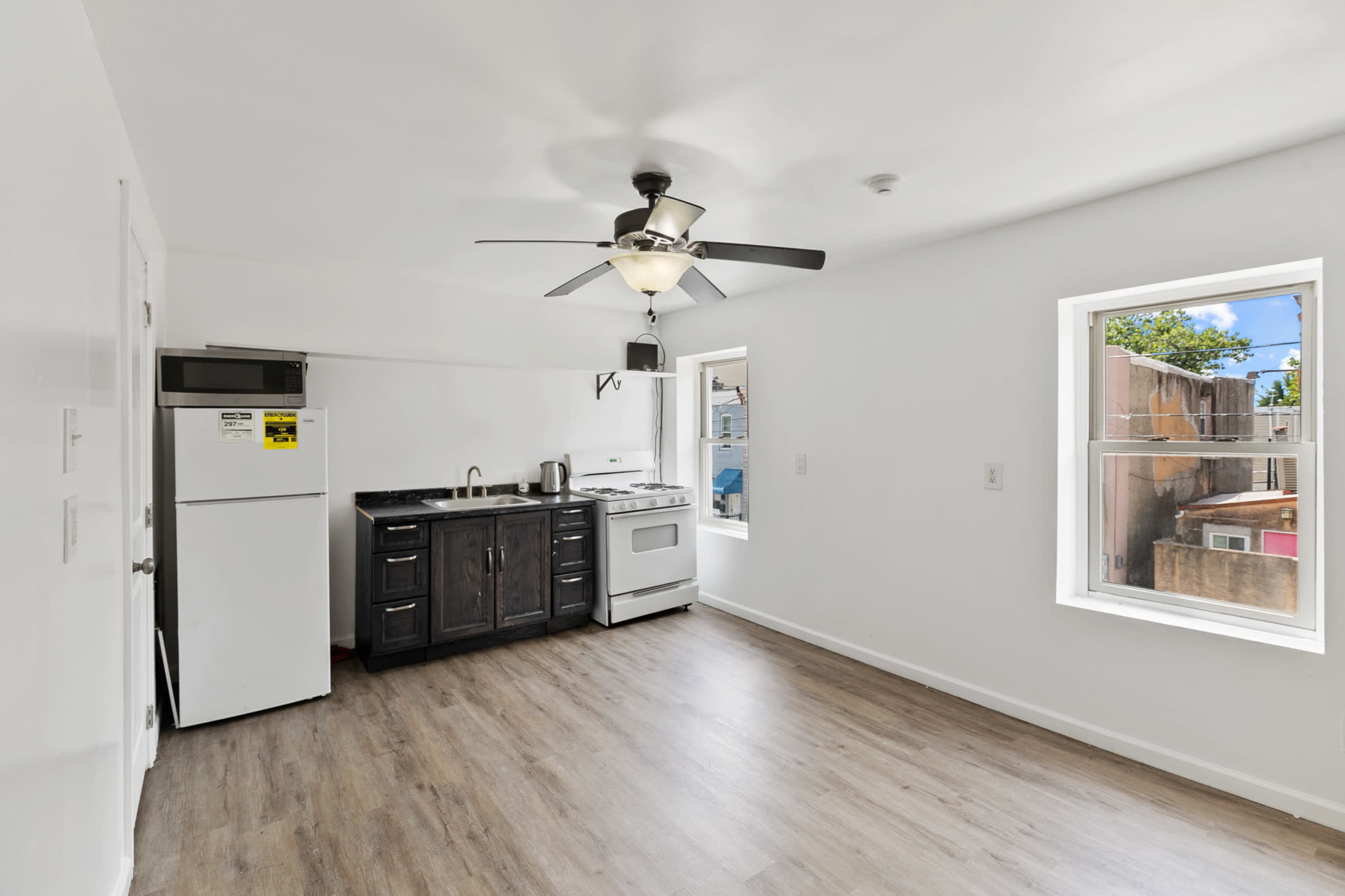 The image shows a bright, empty kitchen space featuring a white ceiling fan, a refrigerator, a stove, and a sink against a white wall with a window.