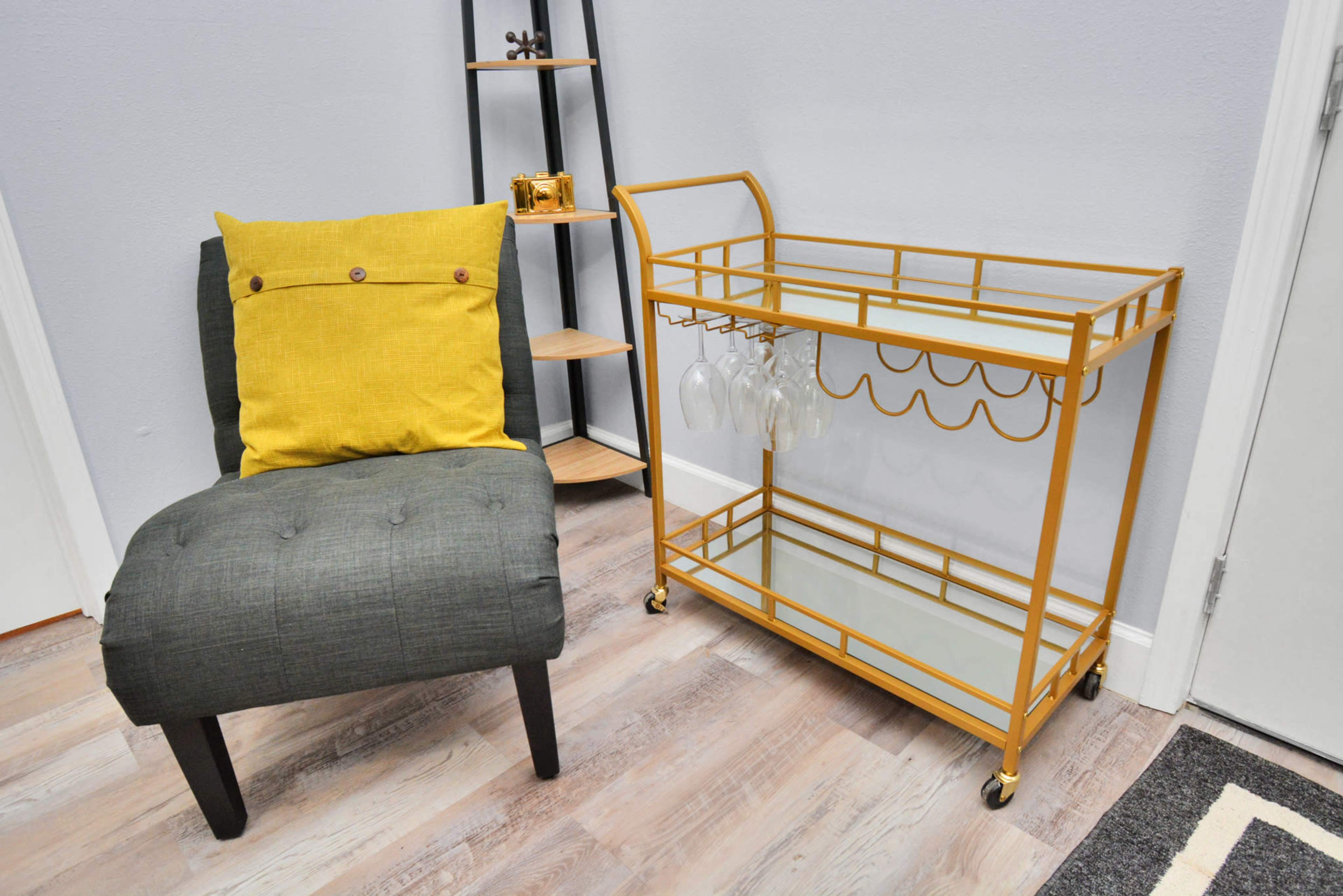 A gray chair with a yellow cushion sits next to a gold bar cart displaying glasses and a small shelf with decorative items.
