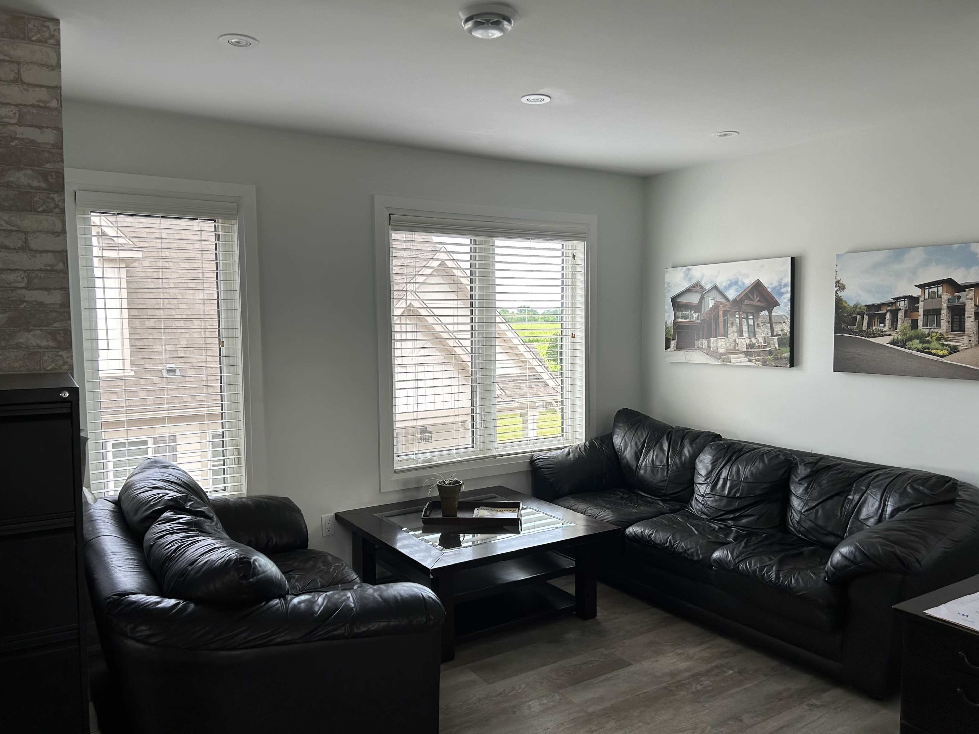 A sitting area features two black leather sofas arranged around a coffee table, with large windows allowing light to enter and framed pictures on the walls.