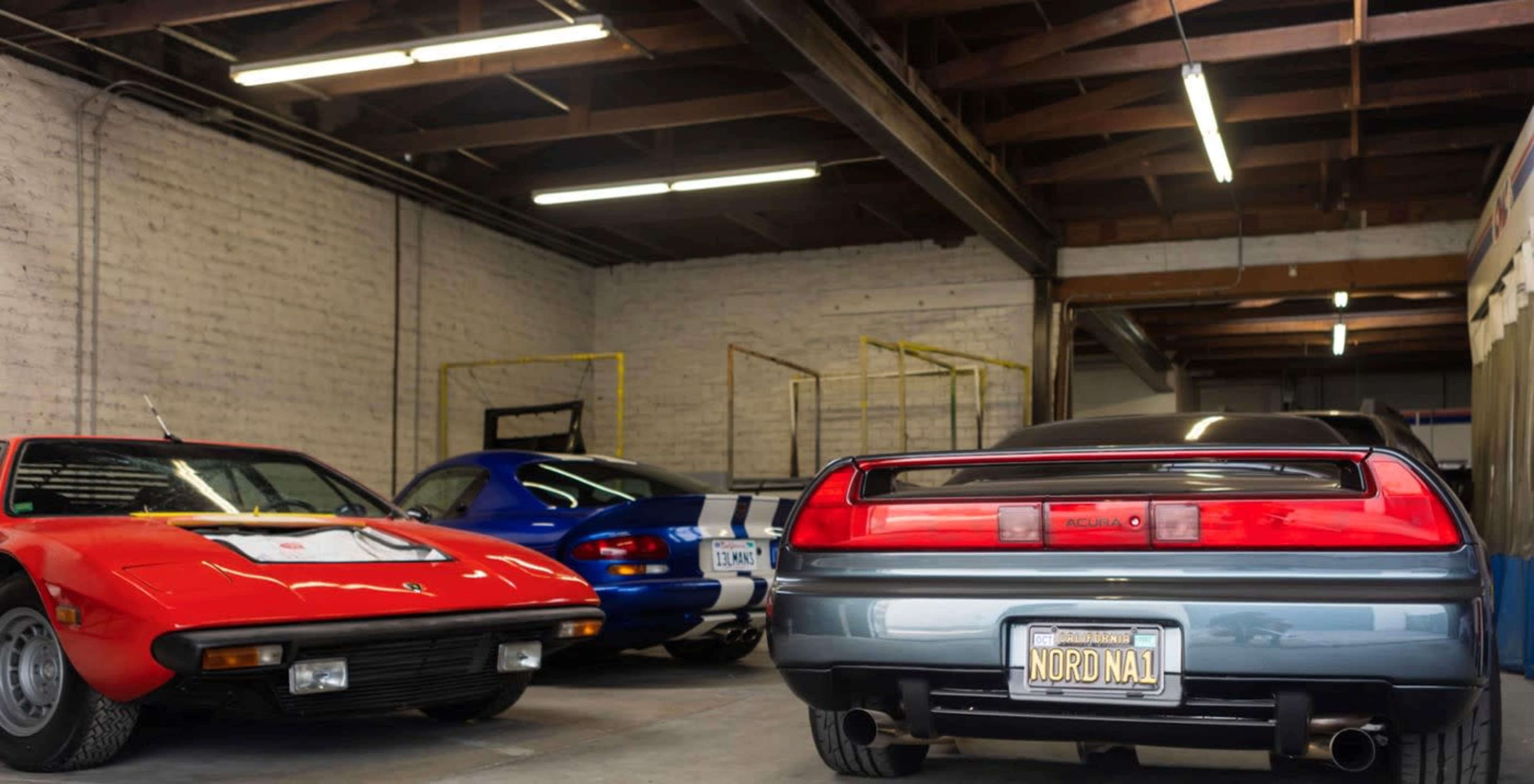 Three classic cars are parked in a garage, featuring a red sports car, a blue coupe, and a green sedan.