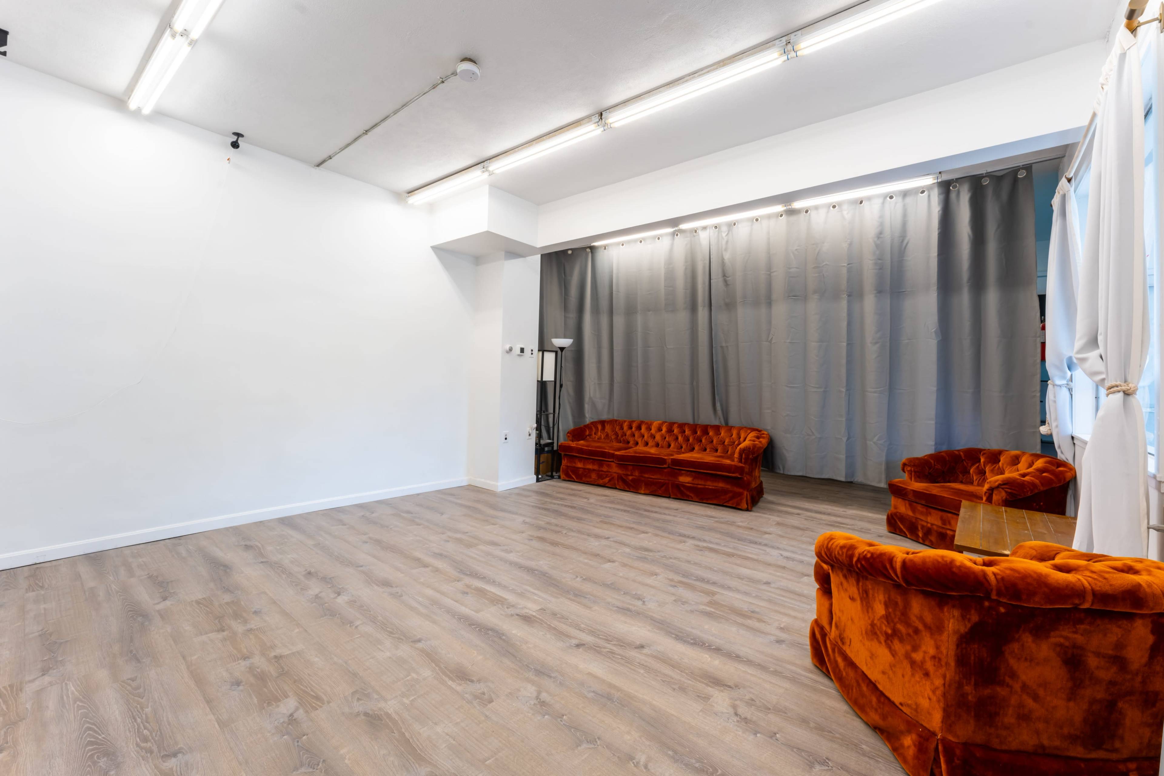 A sparsely furnished room with wooden flooring, featuring two orange velvet sofas and gray curtains along one wall.
