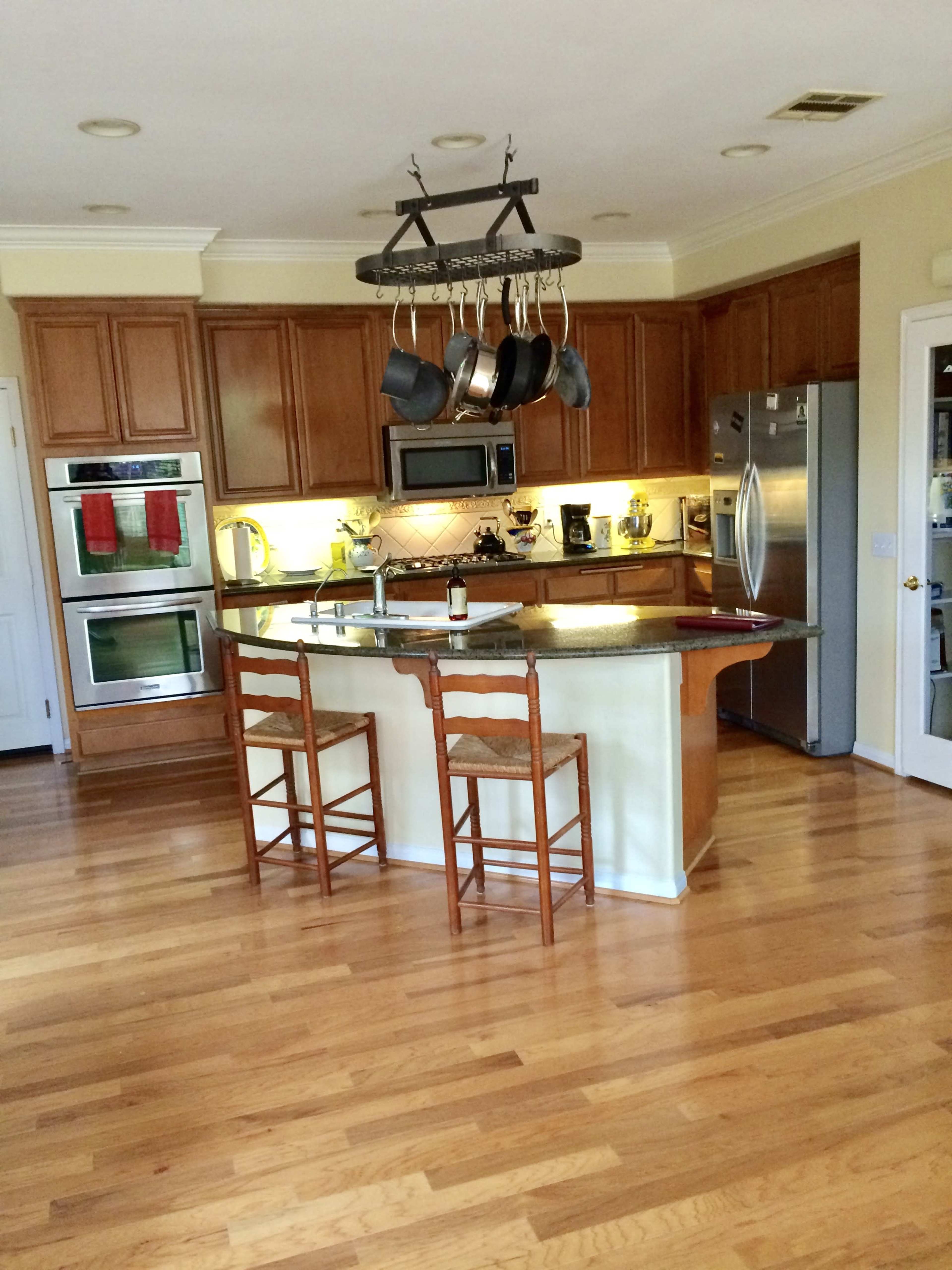 A modern kitchen features wooden cabinets, stainless steel appliances, a central island with two wooden stools, and a pot rack hanging overhead.