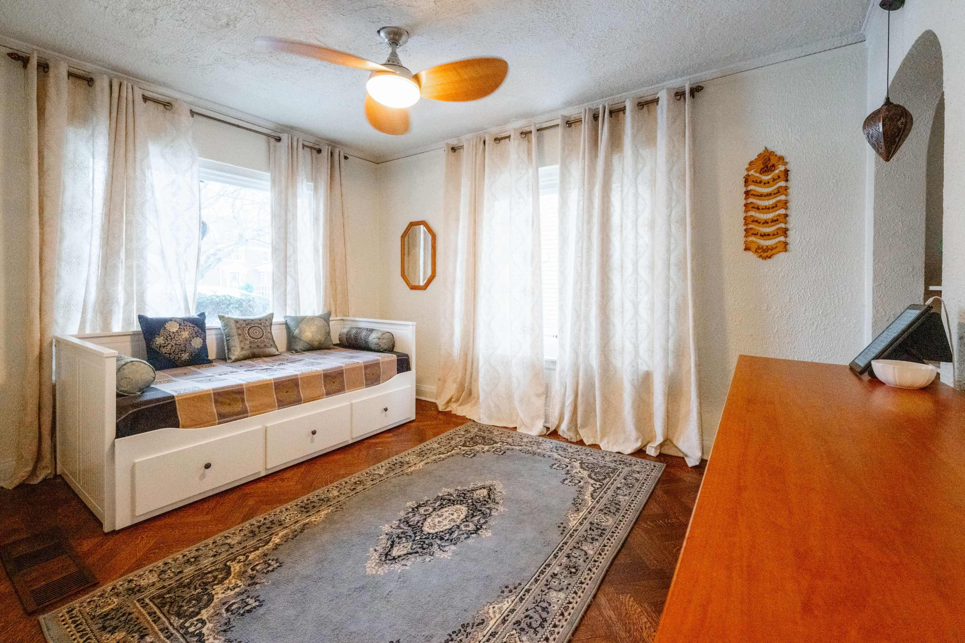 The image shows a cozy room featuring a daybed with decorative pillows, a wooden dresser, a large area rug, and light, flowing curtains over a window.