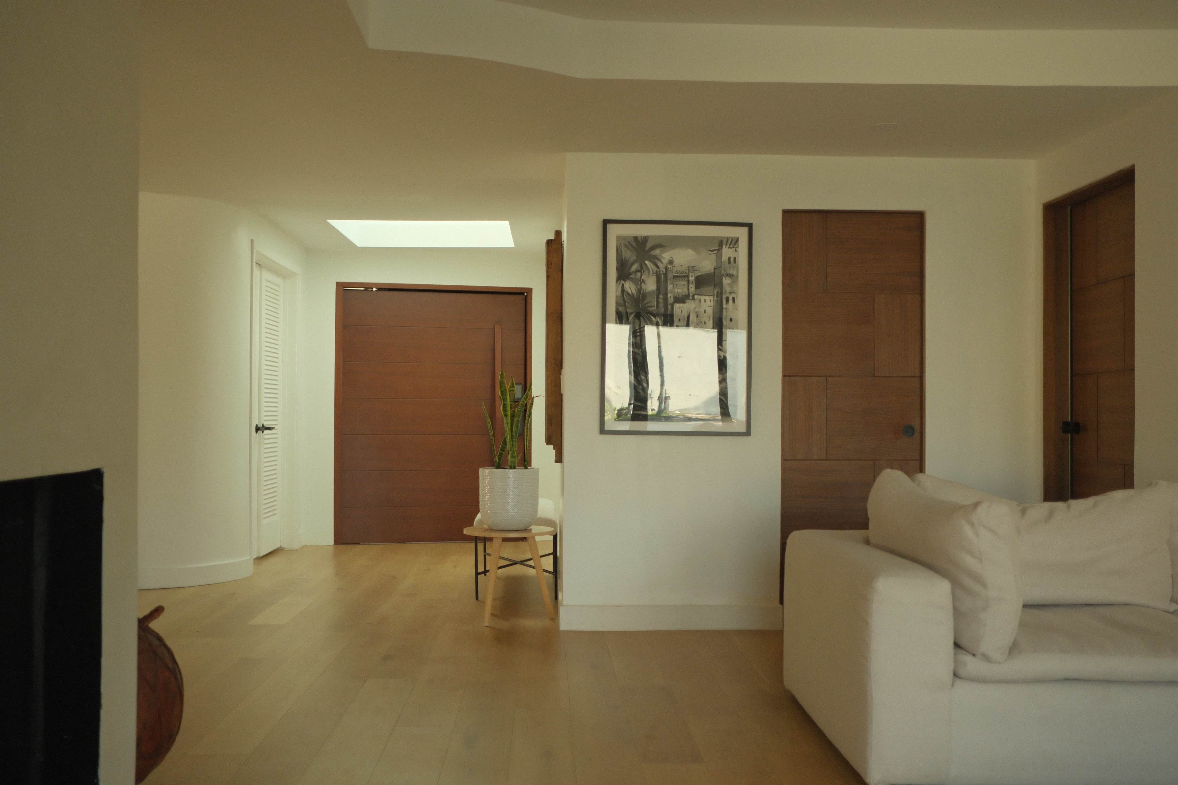 The image shows a minimalist interior space with wooden doors, a framed picture on the wall, a small potted plant, and a white sofa.