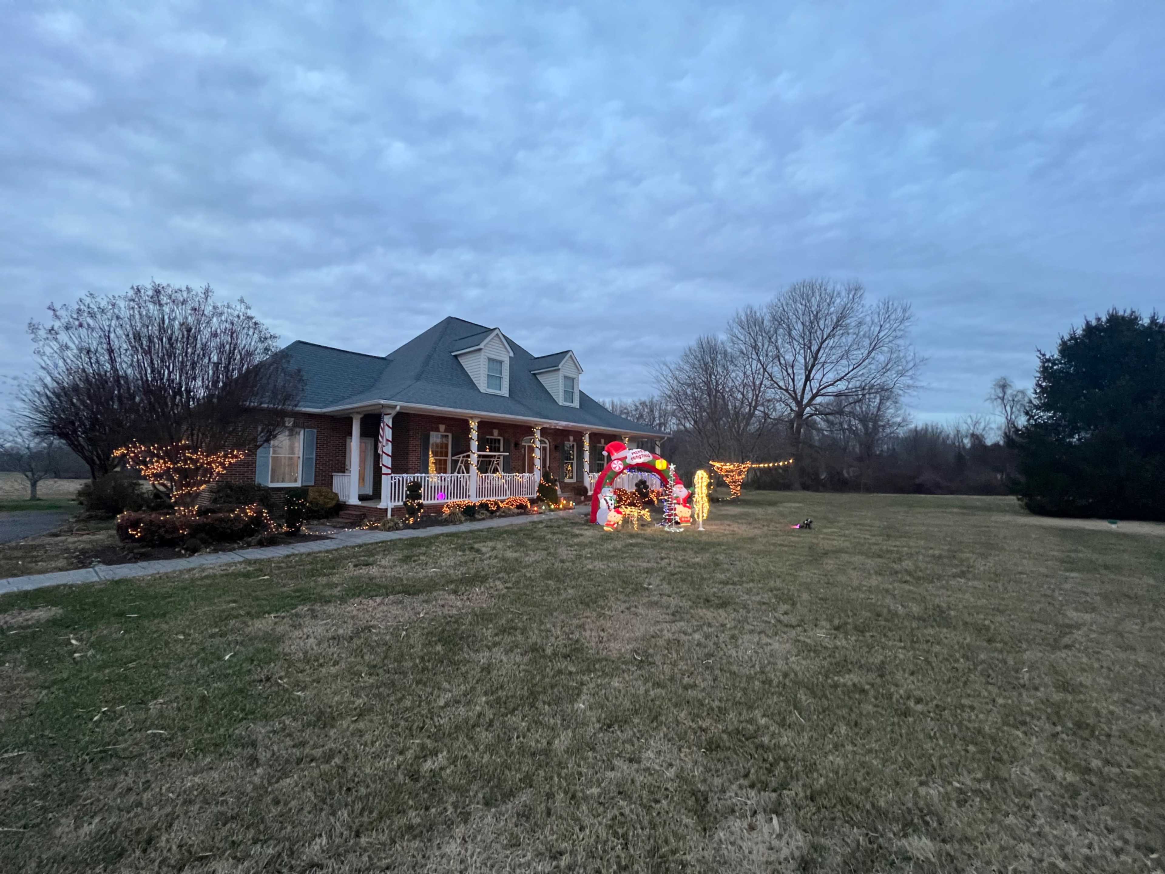 A house is decorated with colorful Christmas lights and inflatable decorations in front of a grassy yard under a cloudy sky.