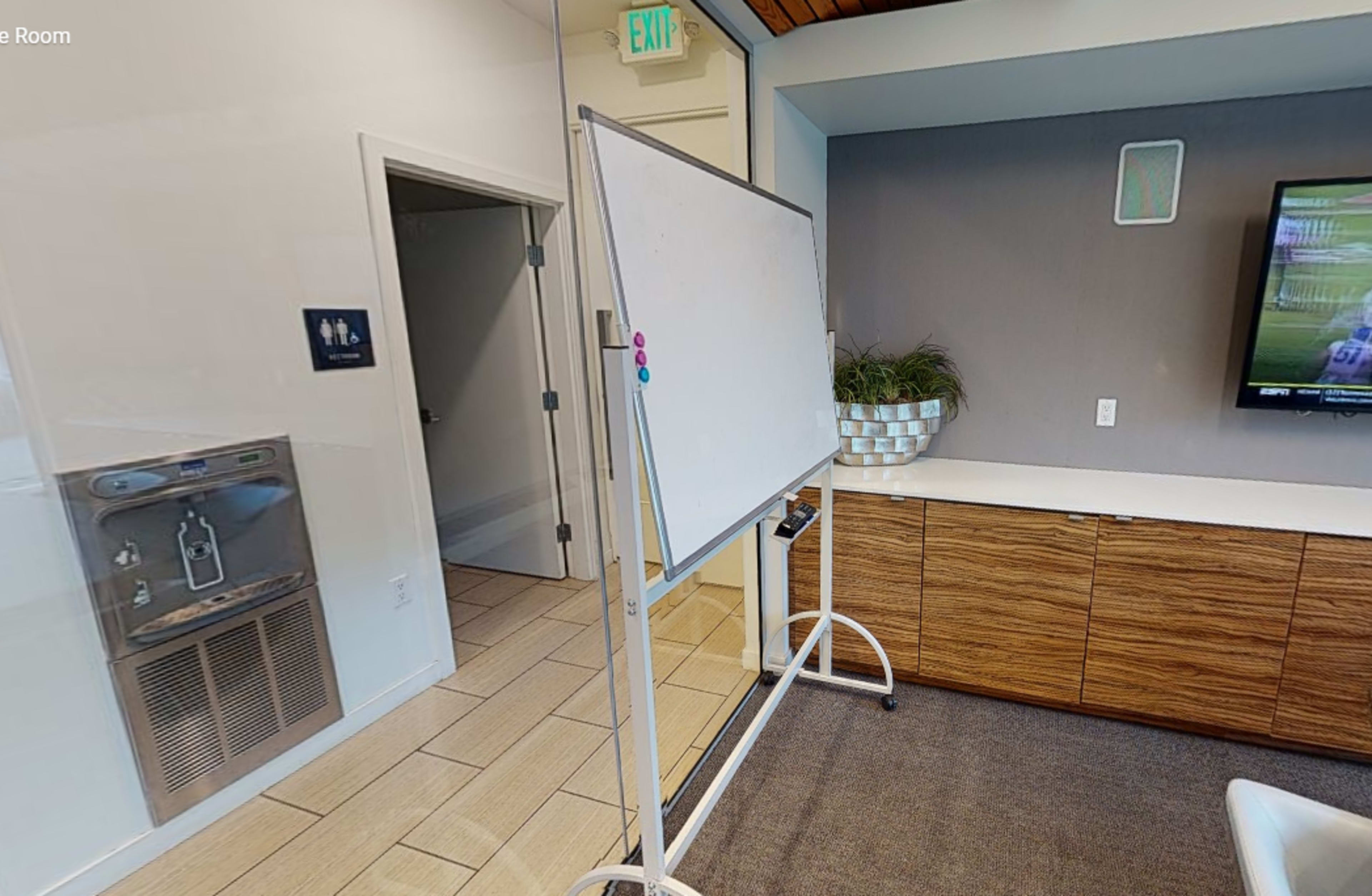A whiteboard on a stand is positioned in a room with a television and wooden cabinetry, alongside a water fountain and a door marked for restrooms.