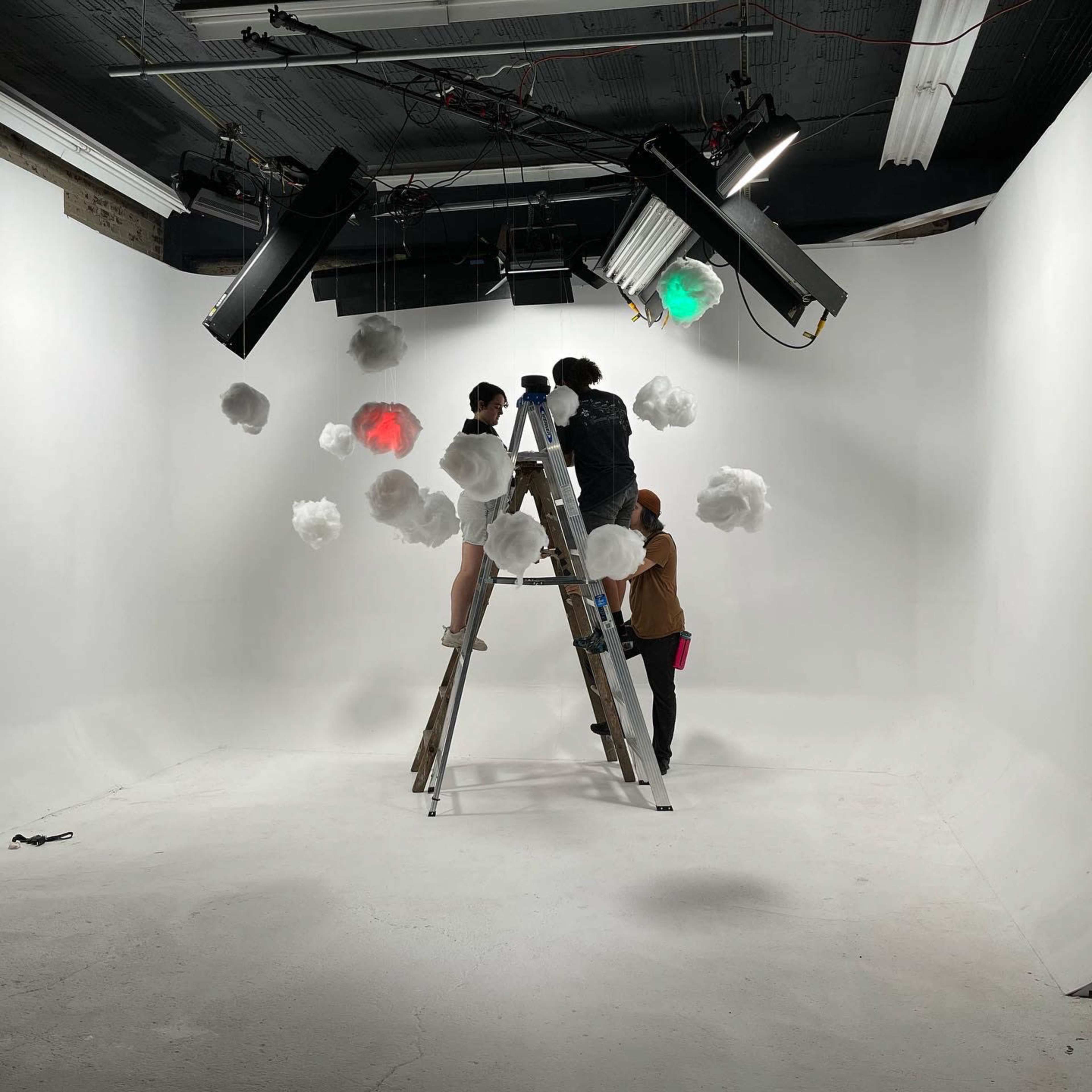 Three people are arranging decorative cloud props on a white studio set using a ladder.