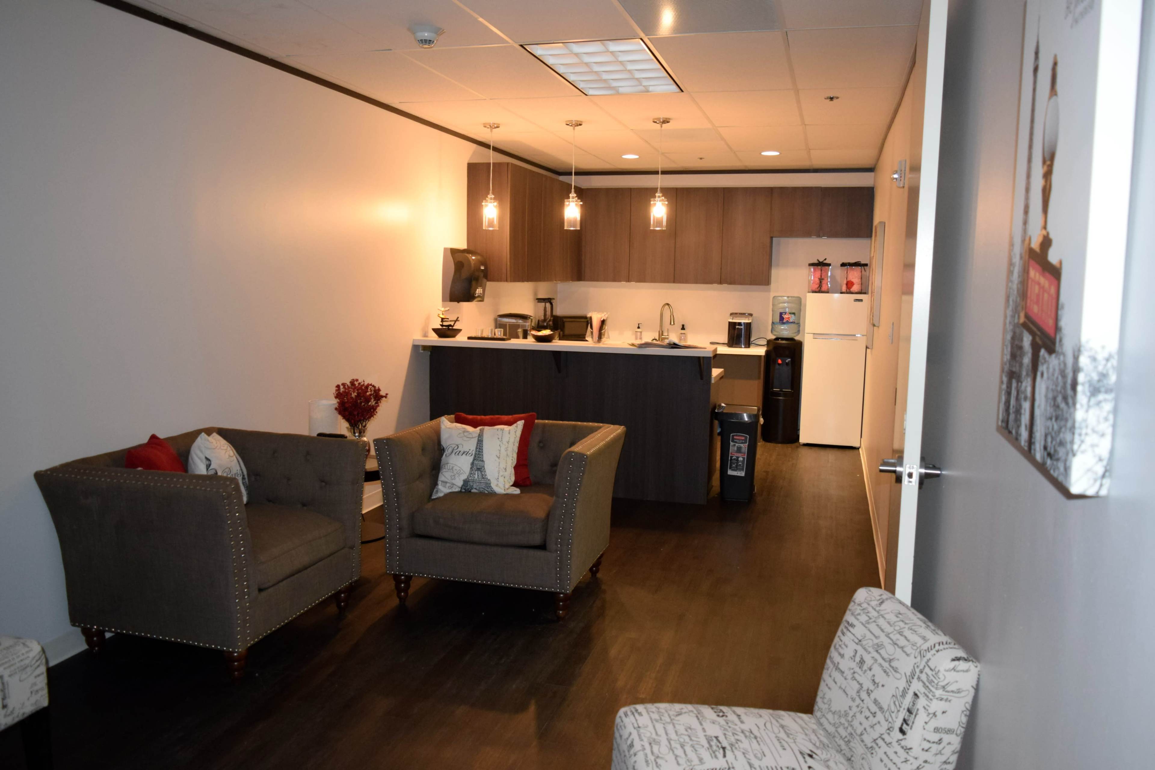 A modern break room with two upholstered chairs, a kitchenette equipped with appliances, and a fridge at the back.