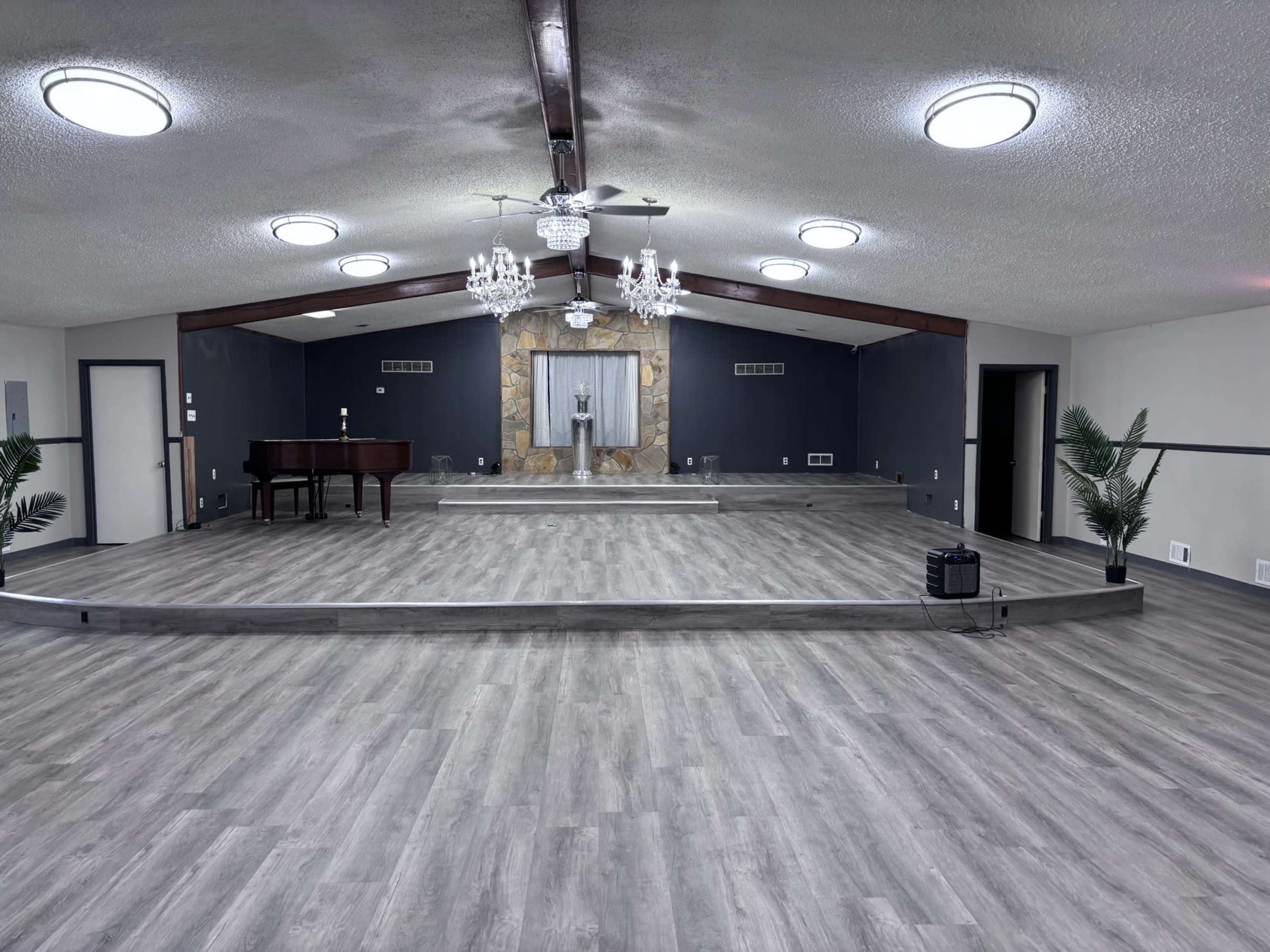 A spacious indoor hall features a raised stage at the back, a piano on the left, and decorative elements, including a chandelier and potted plants.