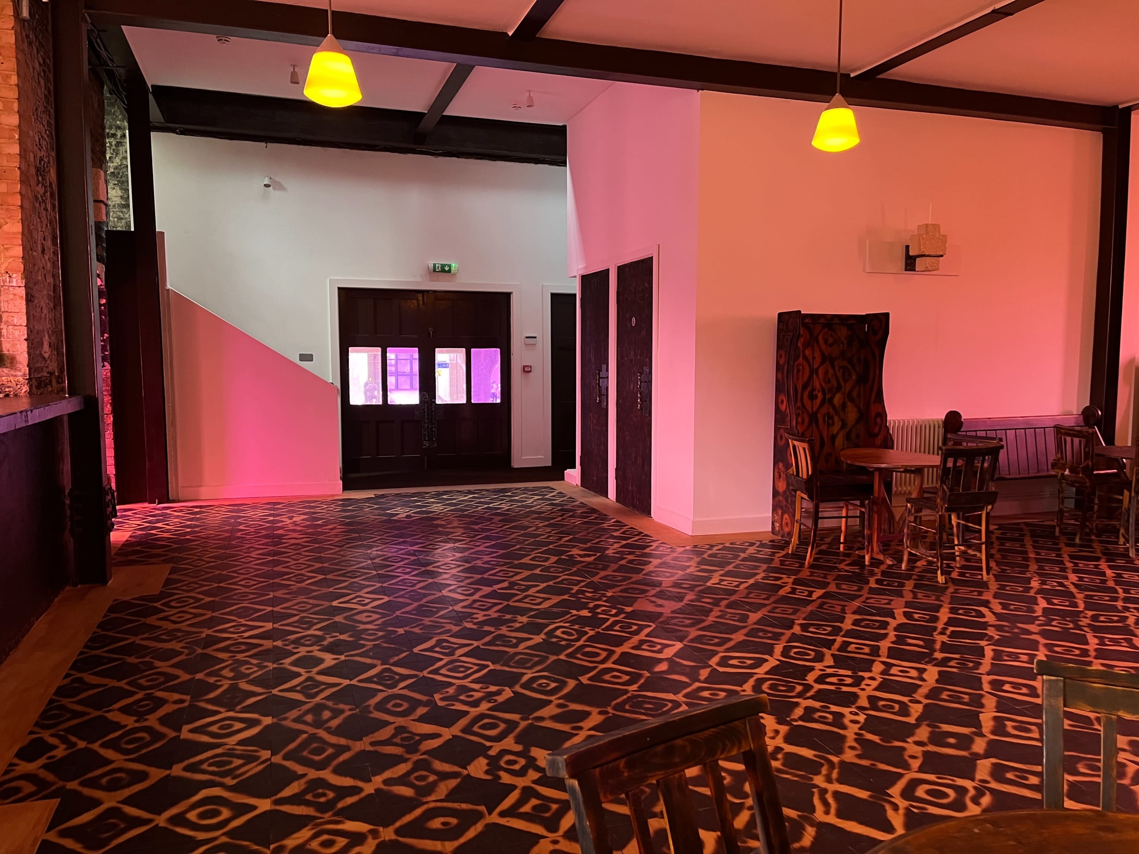 The image showcases an empty room with patterned flooring, featuring a few wooden chairs around tables and a doorway at the far end illuminated by soft pink lighting.