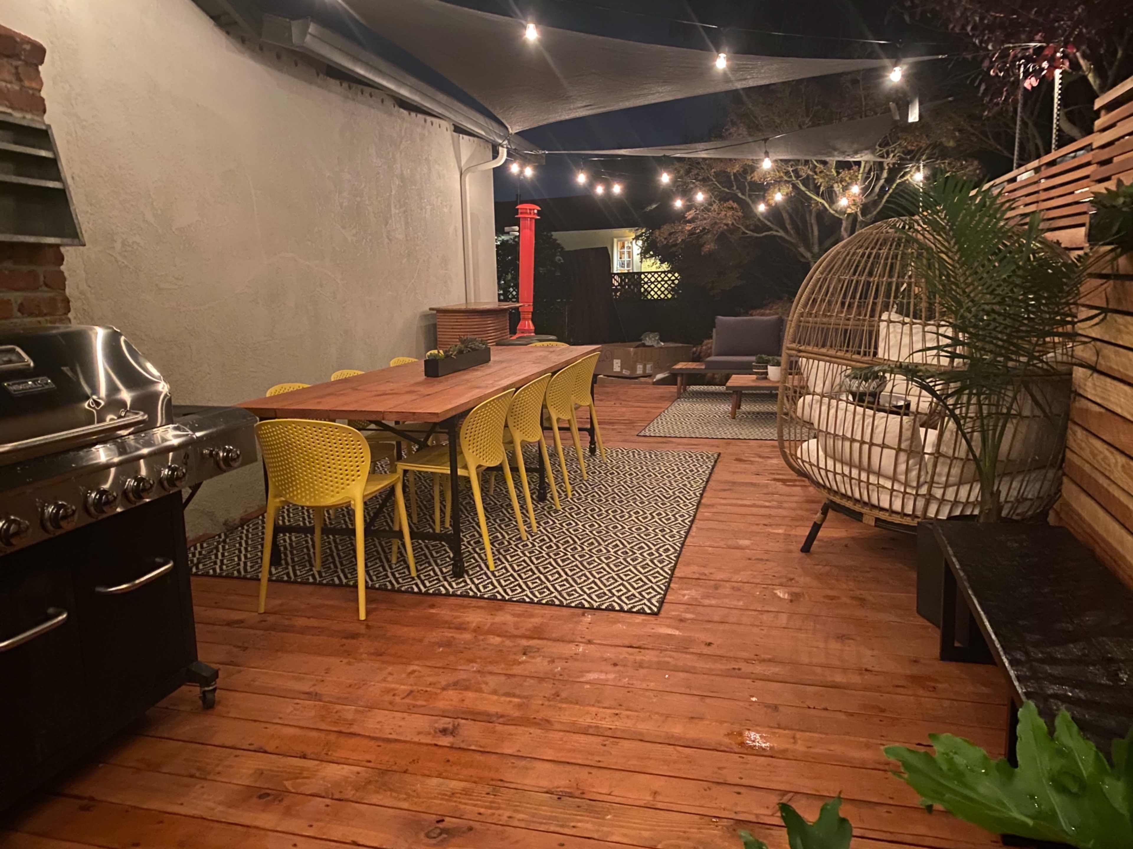 A patio with a long wooden dining table surrounded by yellow chairs, a grill on one side, and hanging string lights illuminating the space at night.