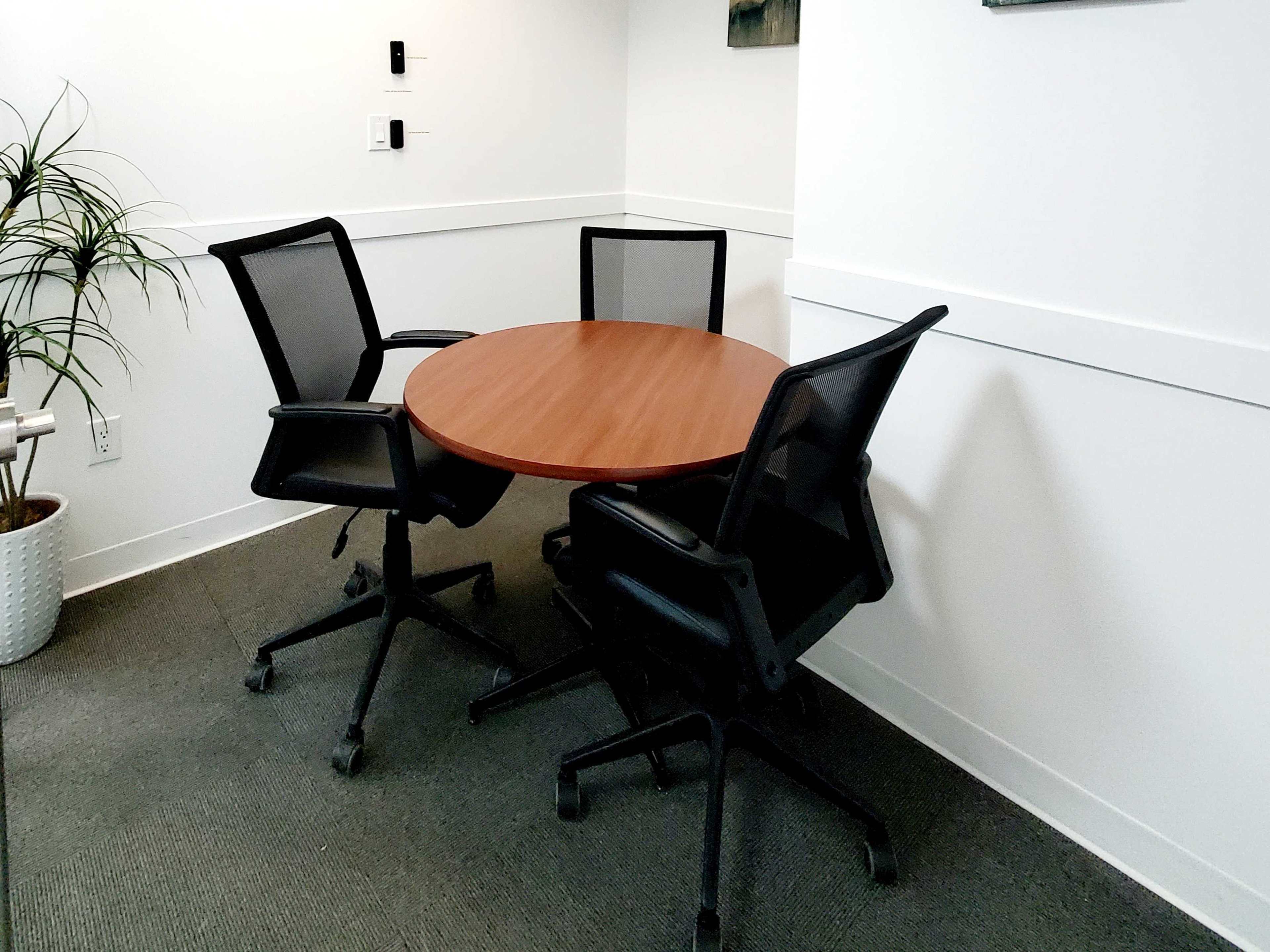 A round wooden table is positioned in a corner of a room with three black mesh chairs around it.