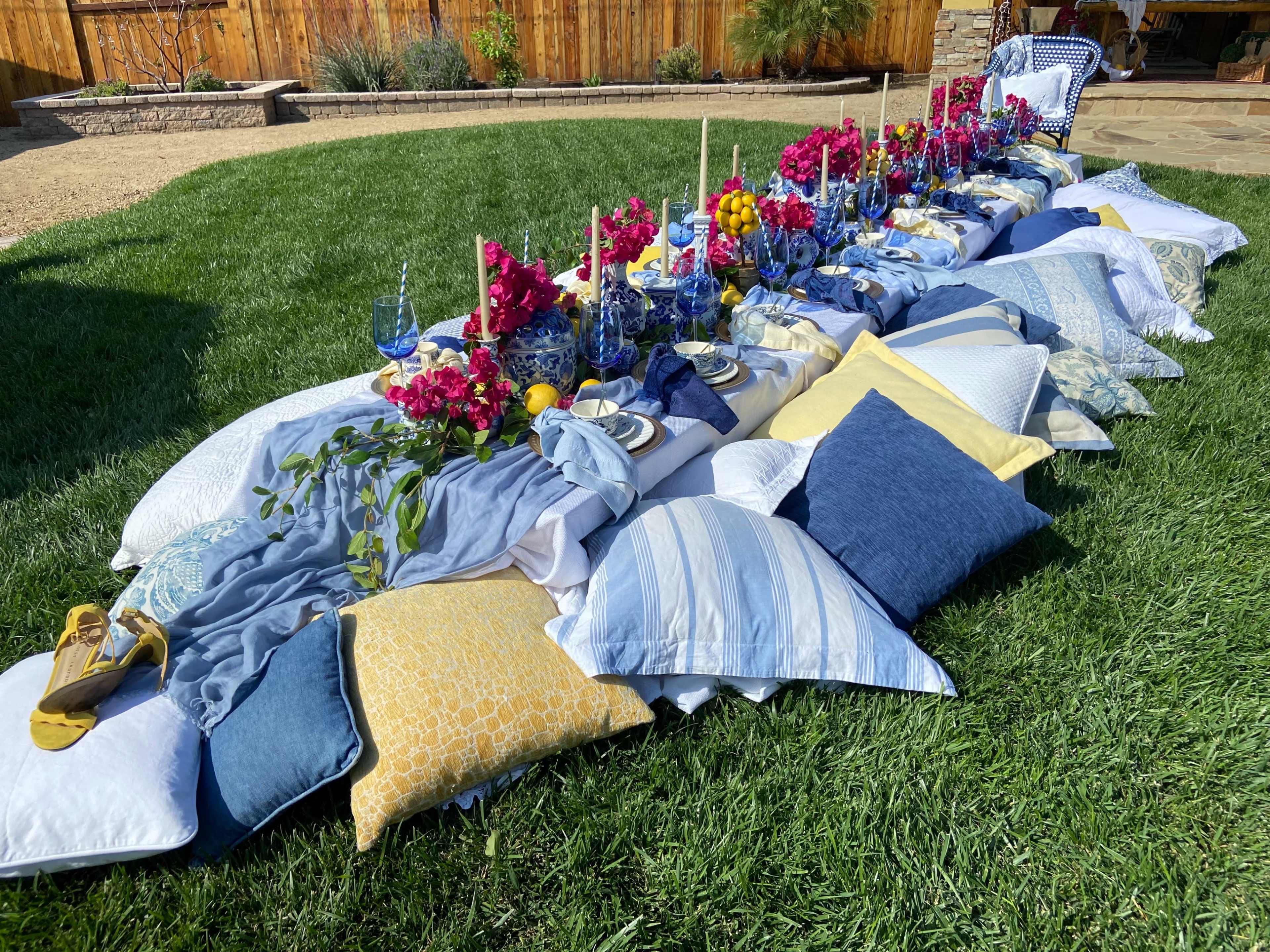 A long table setting with blue and yellow pillows is arranged on a grassy lawn, featuring floral decorations and colorful dishes.