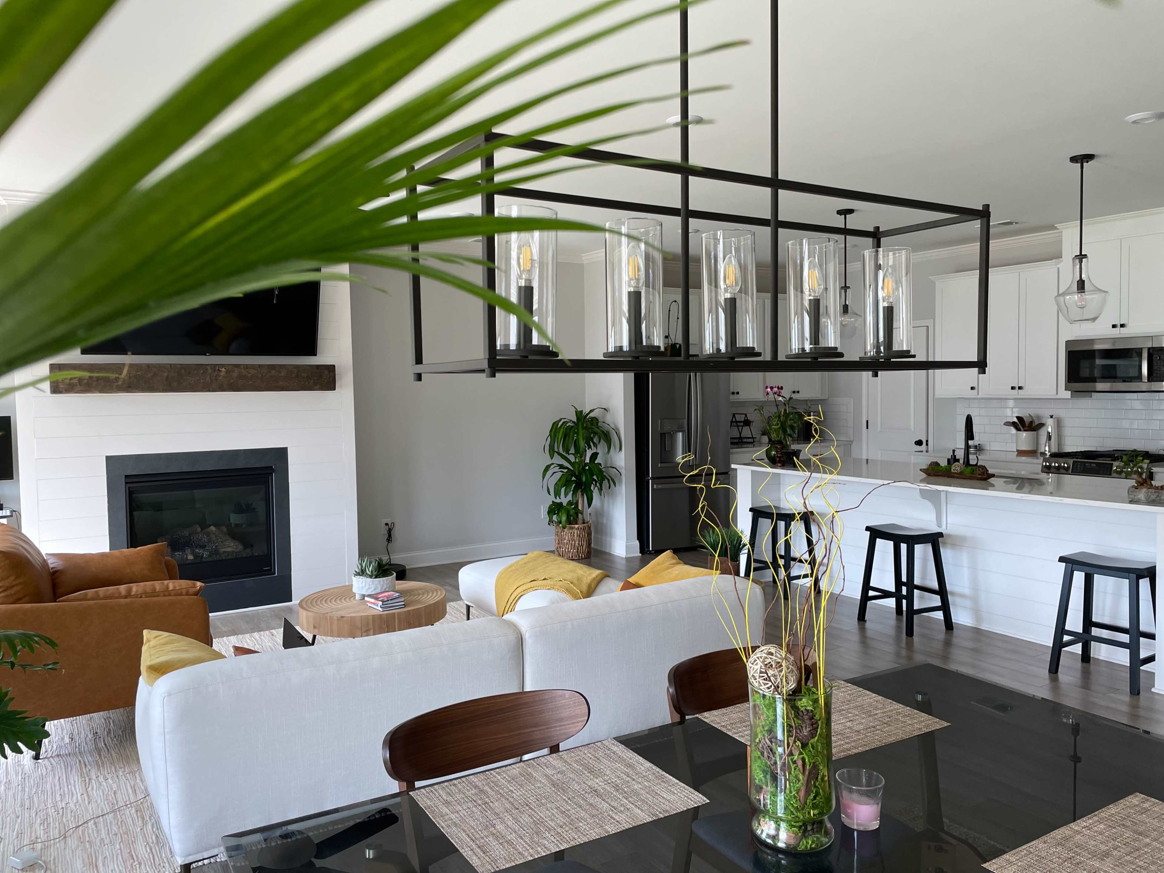 A modern living and dining area features a large chandelier above a dark table, with a beige sofa, brown leather chairs, and a fireplace in the background.
