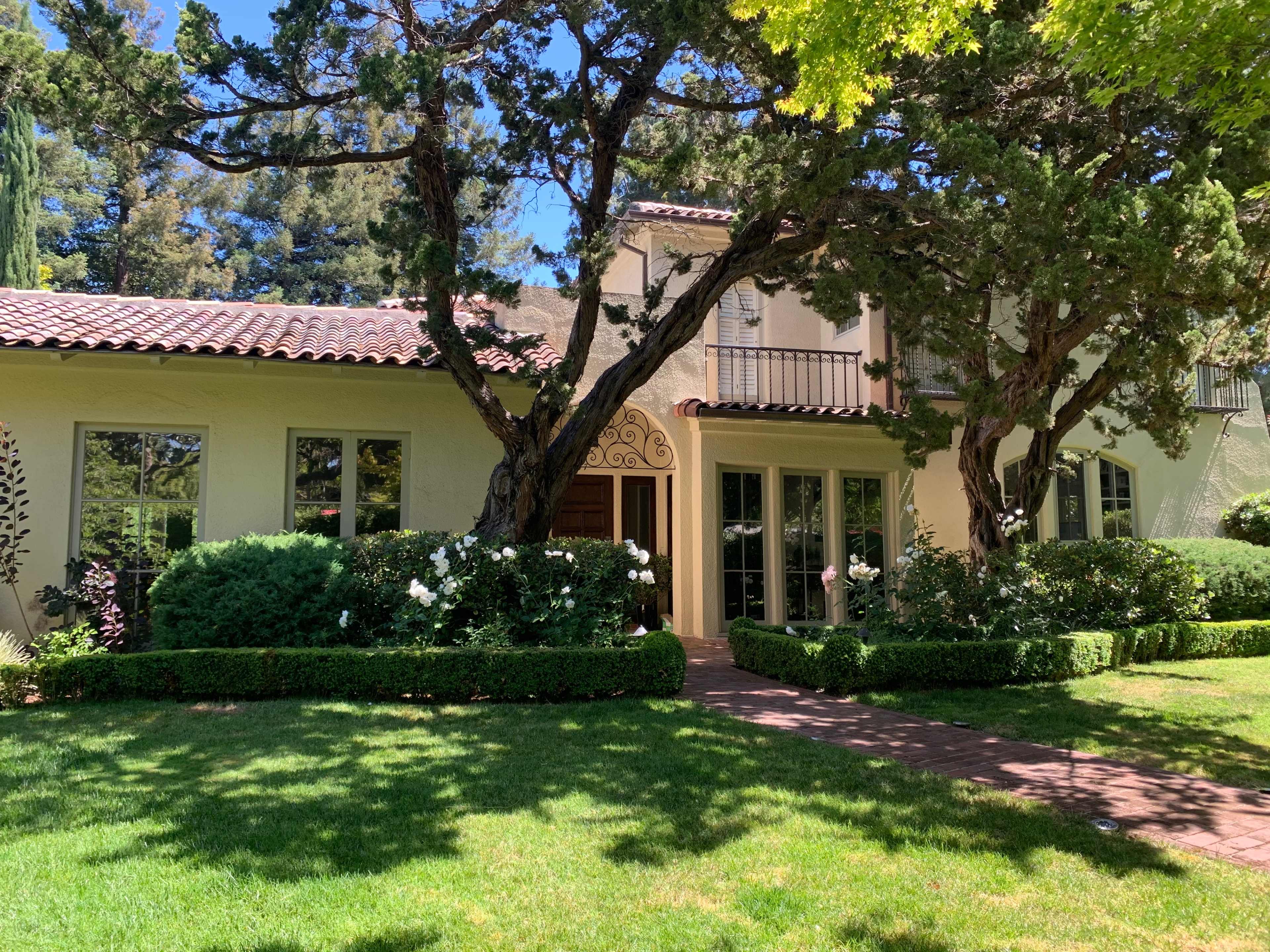 The image shows a two-story house with a tiled roof, surrounded by neatly trimmed hedges and a lawn, under a clear blue sky.
