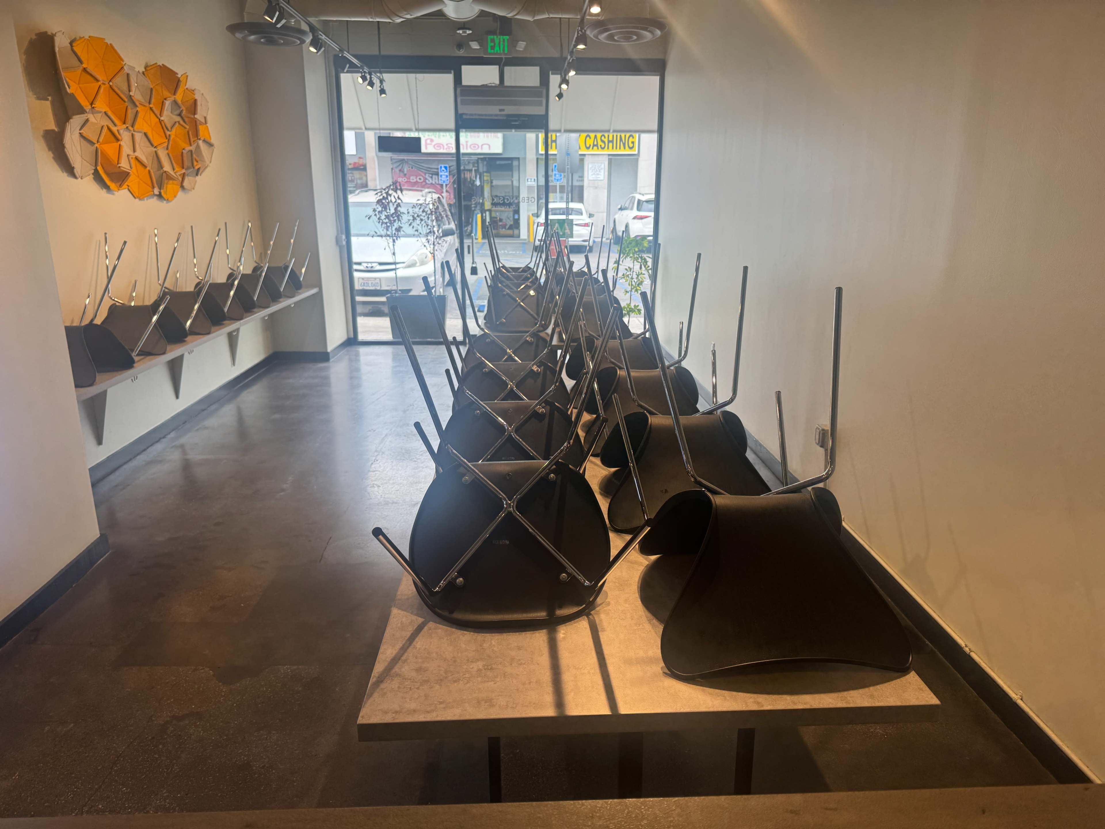 The image shows a café interior with tables that are stacked upside down on top of each other and empty chairs placed against the walls.