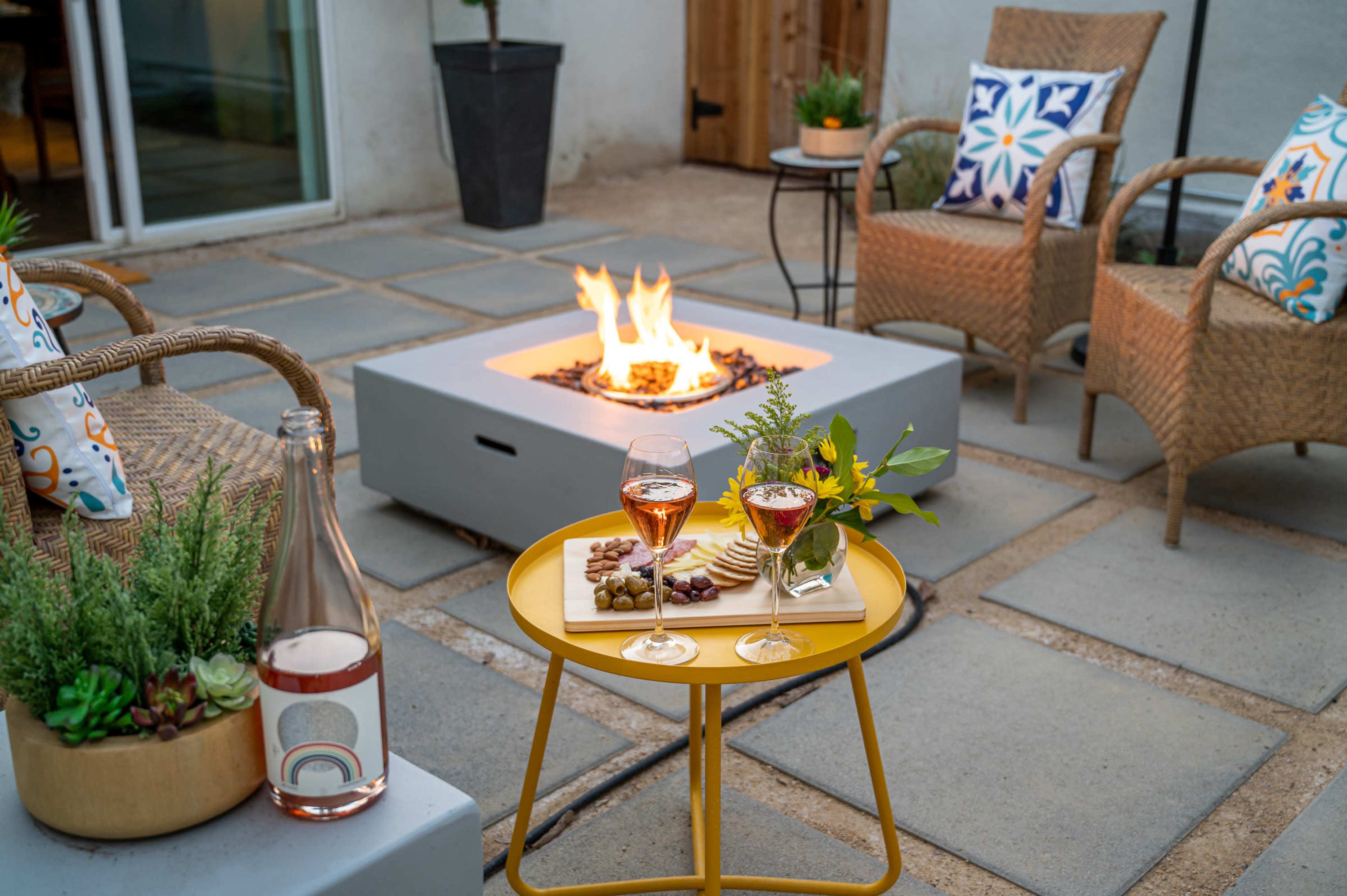 A cozy outdoor patio features a fire pit surrounded by two wicker chairs, with a small table holding wine glasses, a bottle, and a platter of snacks.