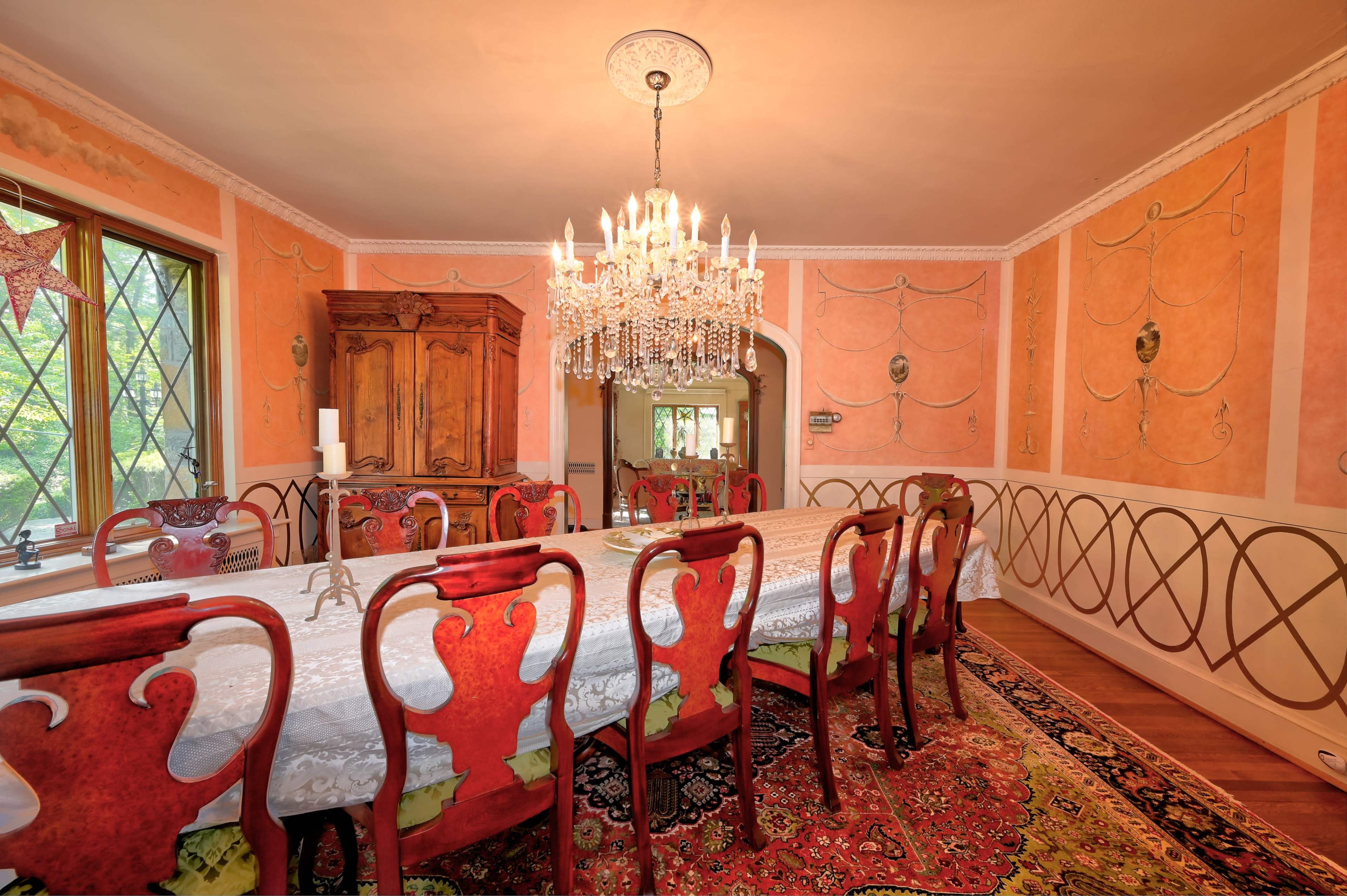 A dining room with a long table surrounded by ornate chairs, a chandelier overhead, and decorative wallpaper featuring a blend of colors and patterns.