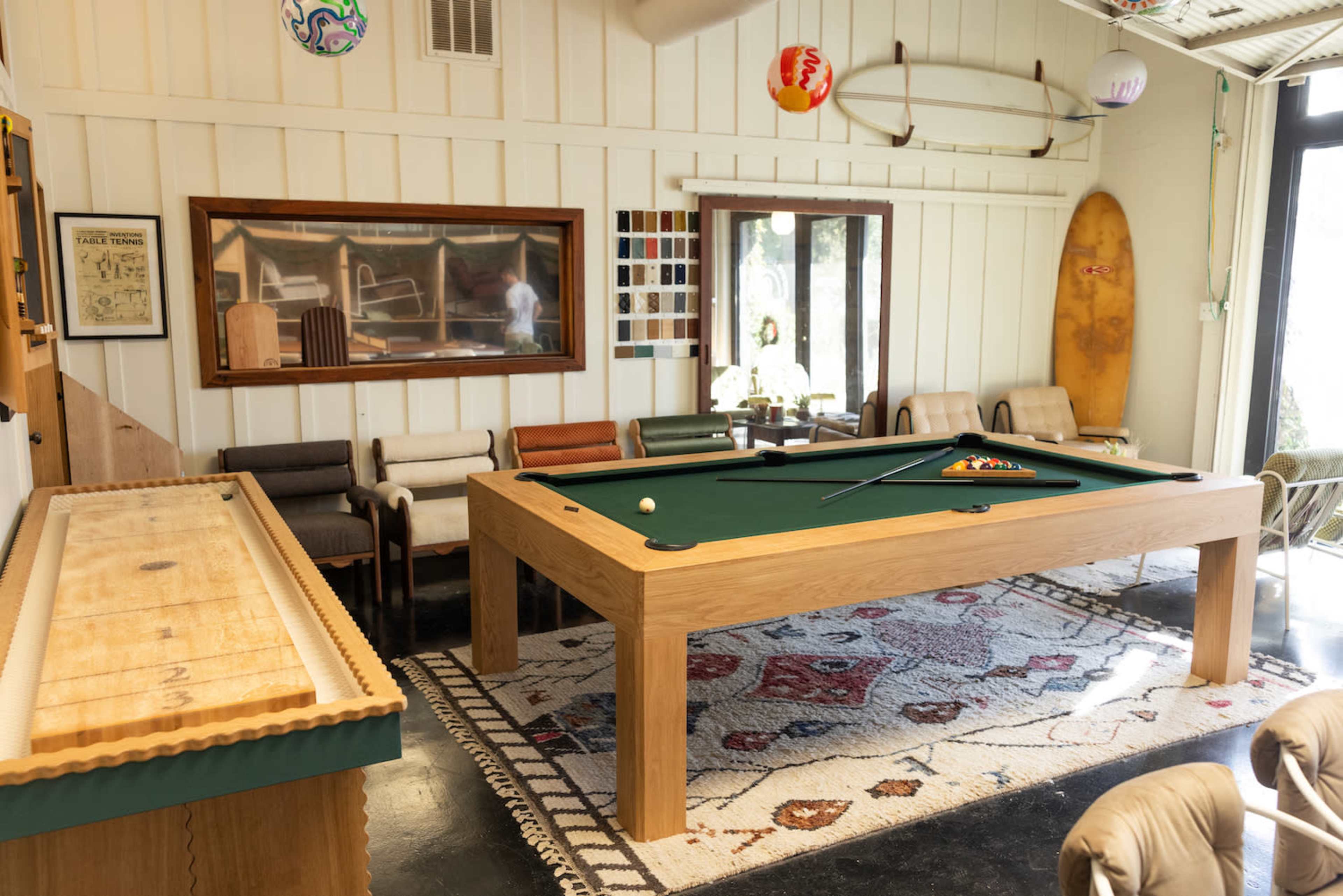 A room with a pool table in the center, surrounded by various seating options and decorative items, including a surfboard and wall art.