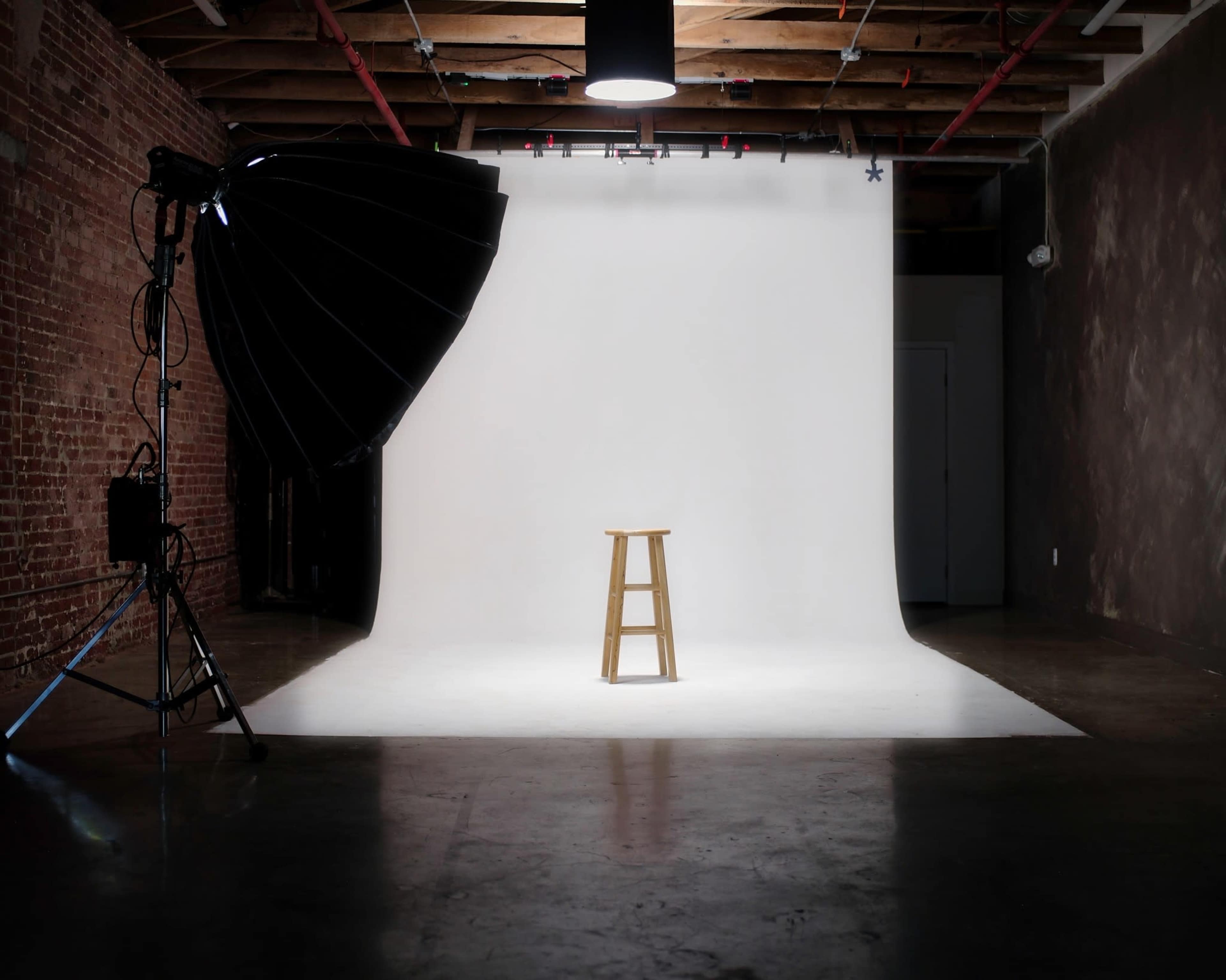 A photography studio features a wooden stool positioned on a white backdrop, illuminated by a large softbox light.