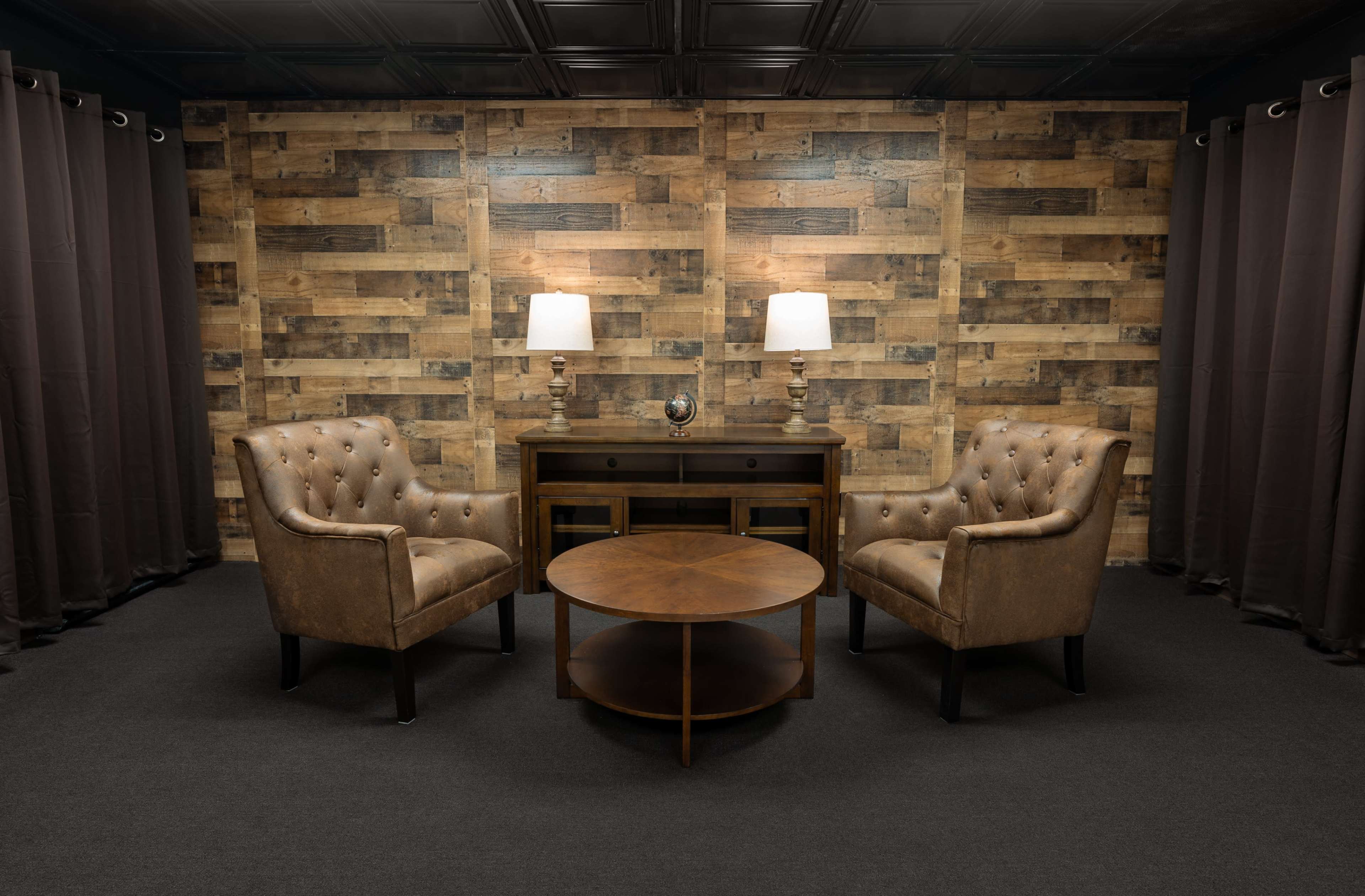 A cozy seating area with two tufted leather chairs facing a round wooden coffee table in front of a wooden wall and dark curtains.
