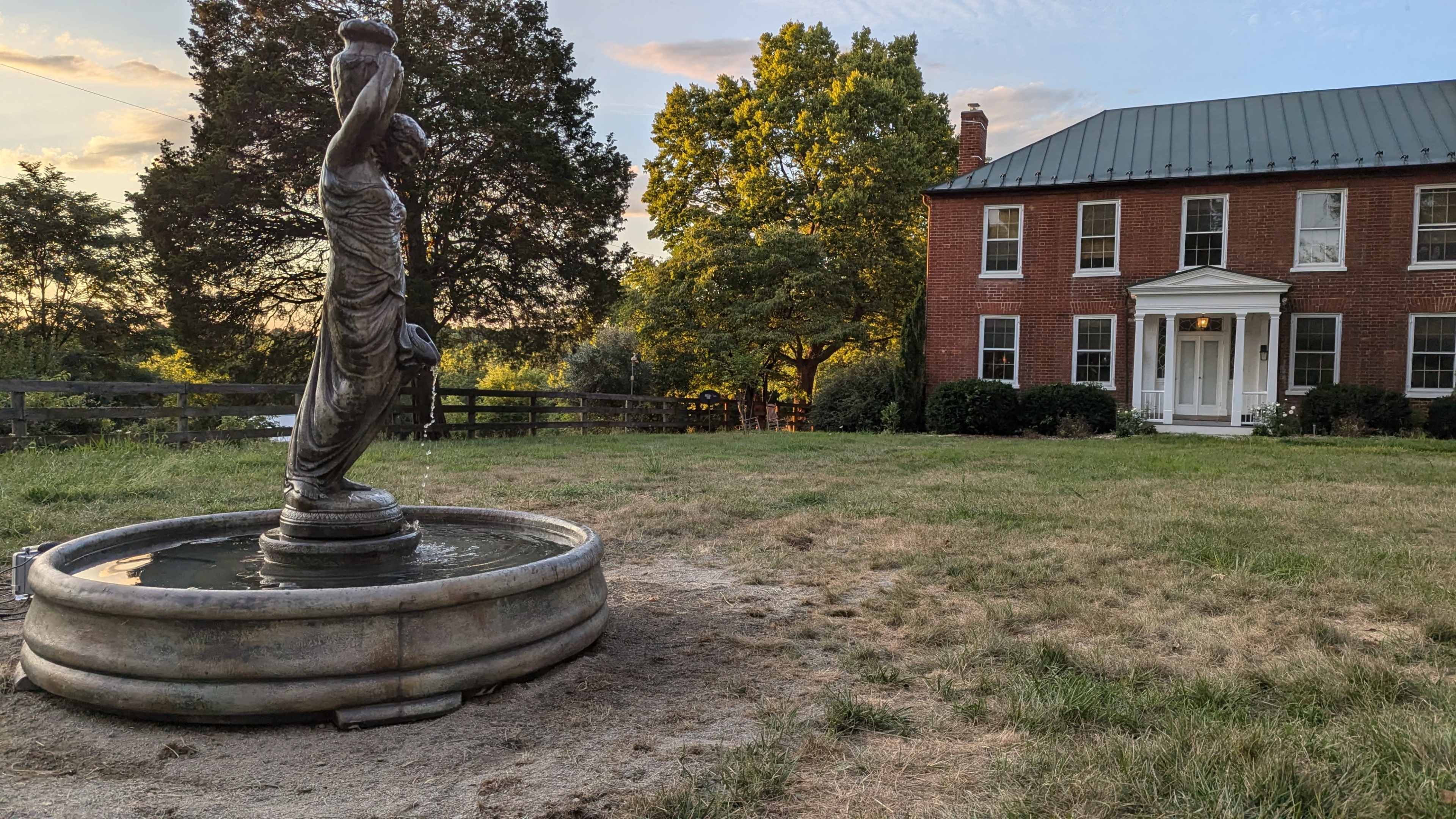 A statue of a woman holding a vessel stands in a fountain on a grassy area in front of a red brick house with a green roof.