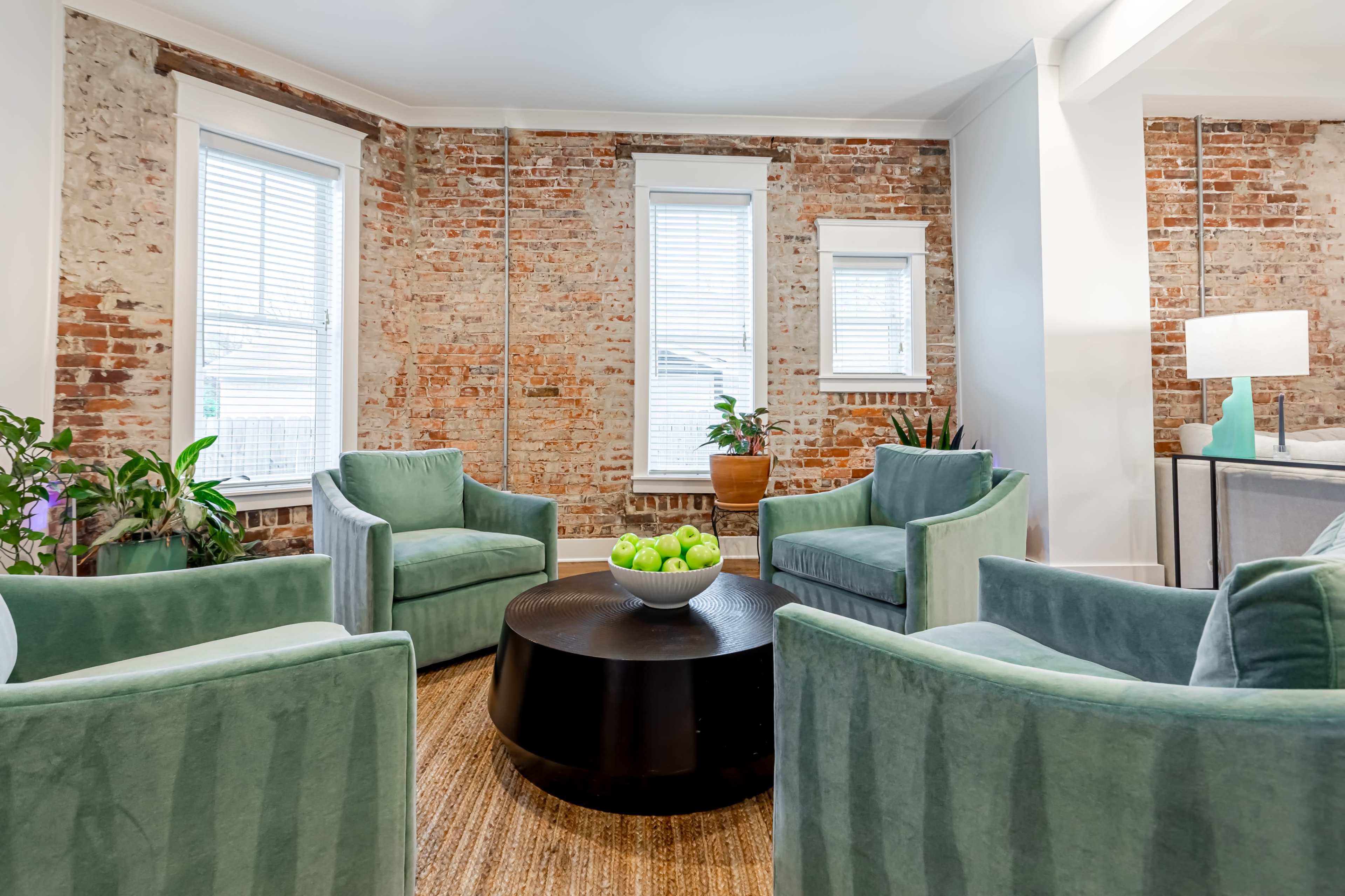 The image shows a seating area with four green upholstered chairs arranged around a round black coffee table, set against exposed brick walls and natural light coming through windows.
