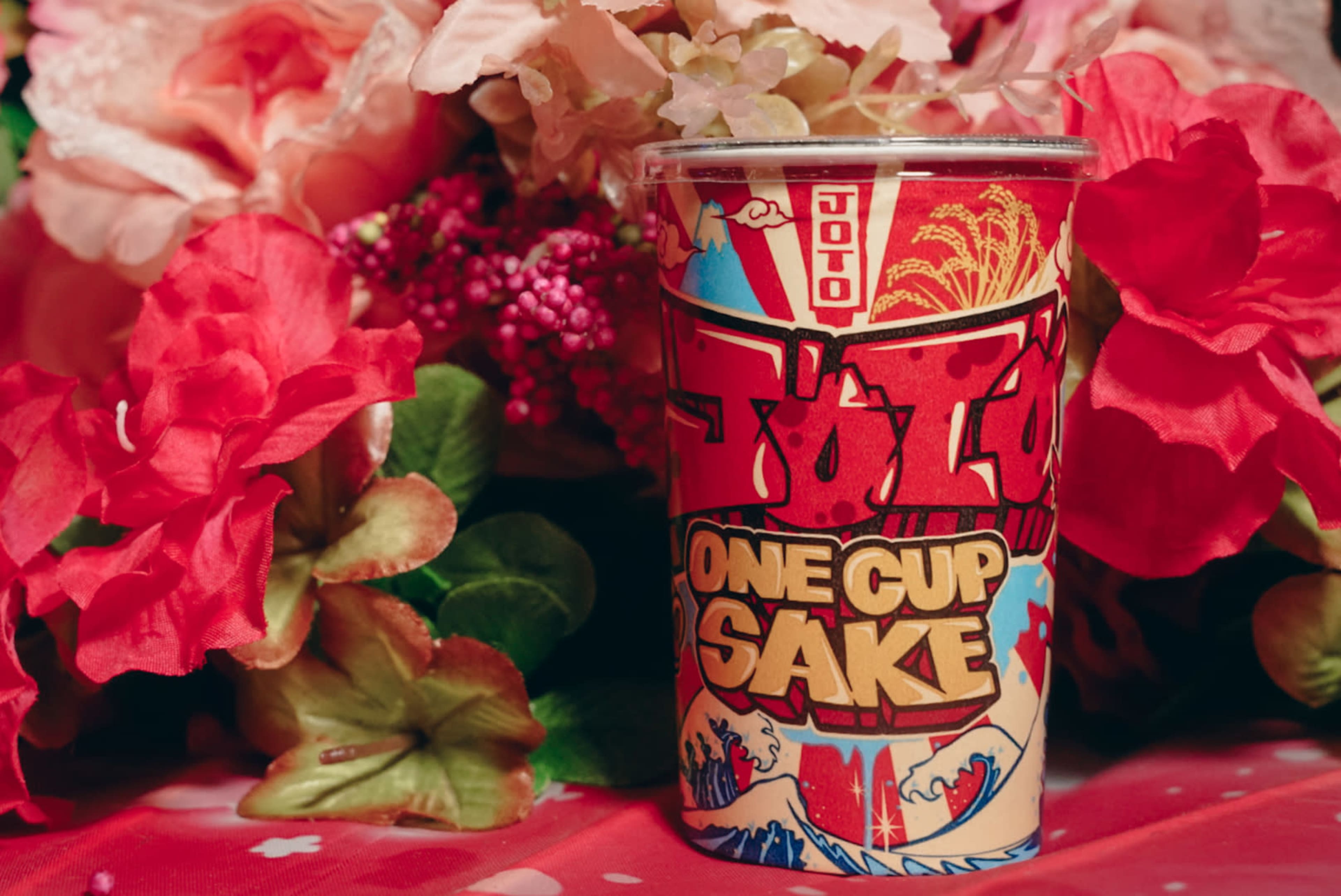 A cup of sake featuring a colorful, artistic design sits among vibrant pink flowers.