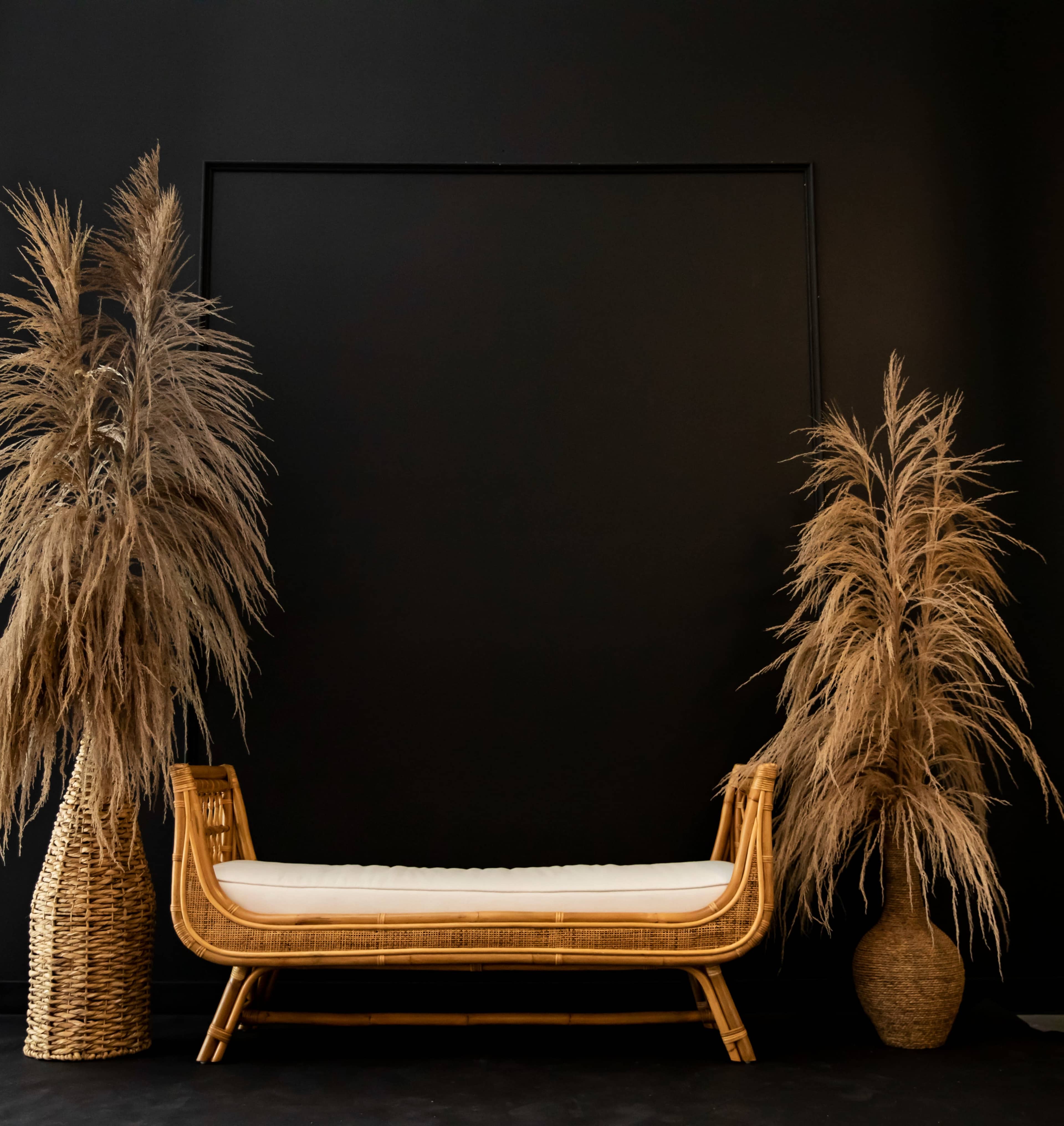The image shows a rattan daybed placed against a black wall, flanked by two tall, dried grass arrangements in woven containers.