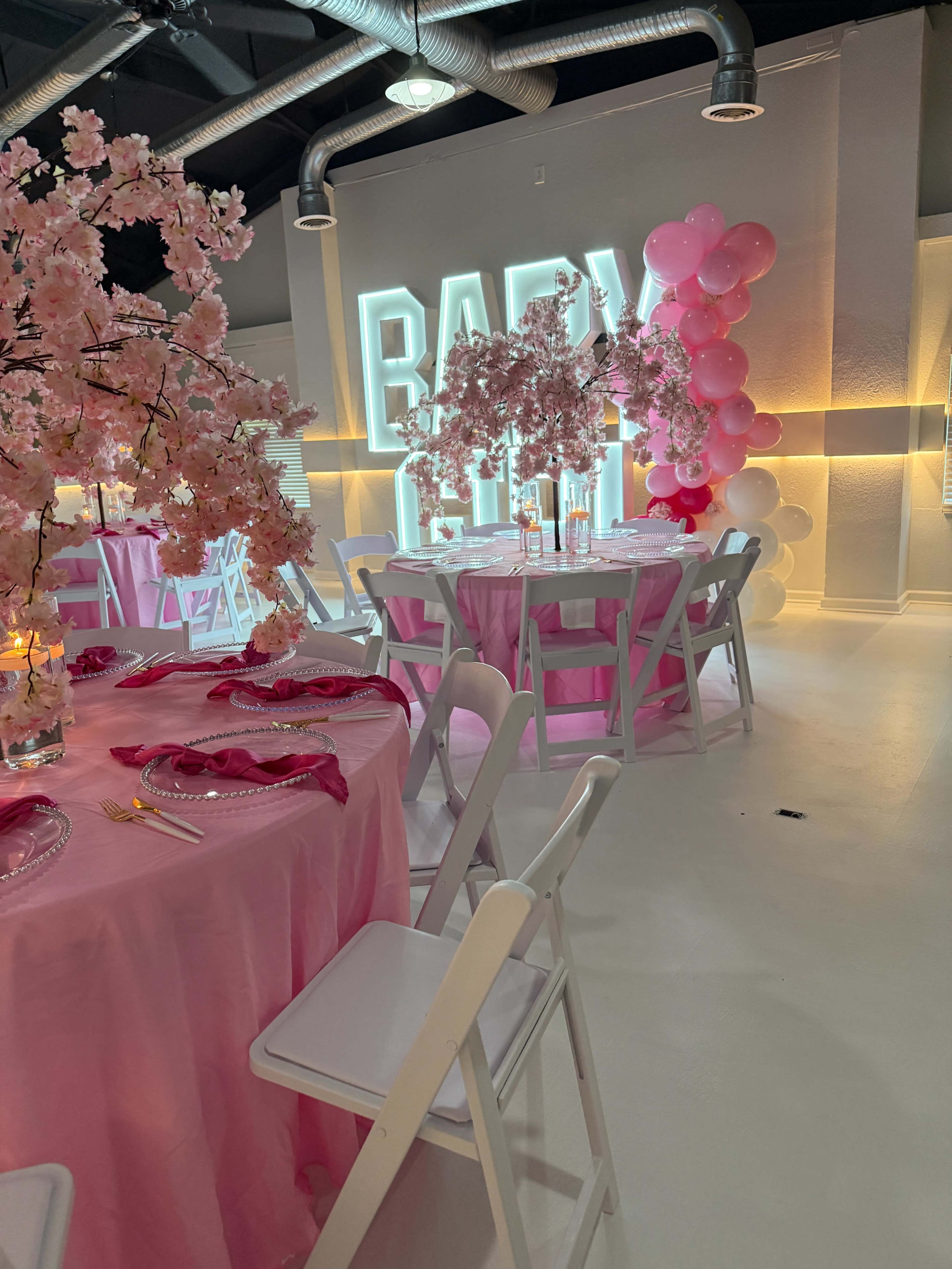 The scene depicts a vibrant event space with pink table settings, cherry blossom centerpieces, and neon "BABY" signage against a white backdrop.