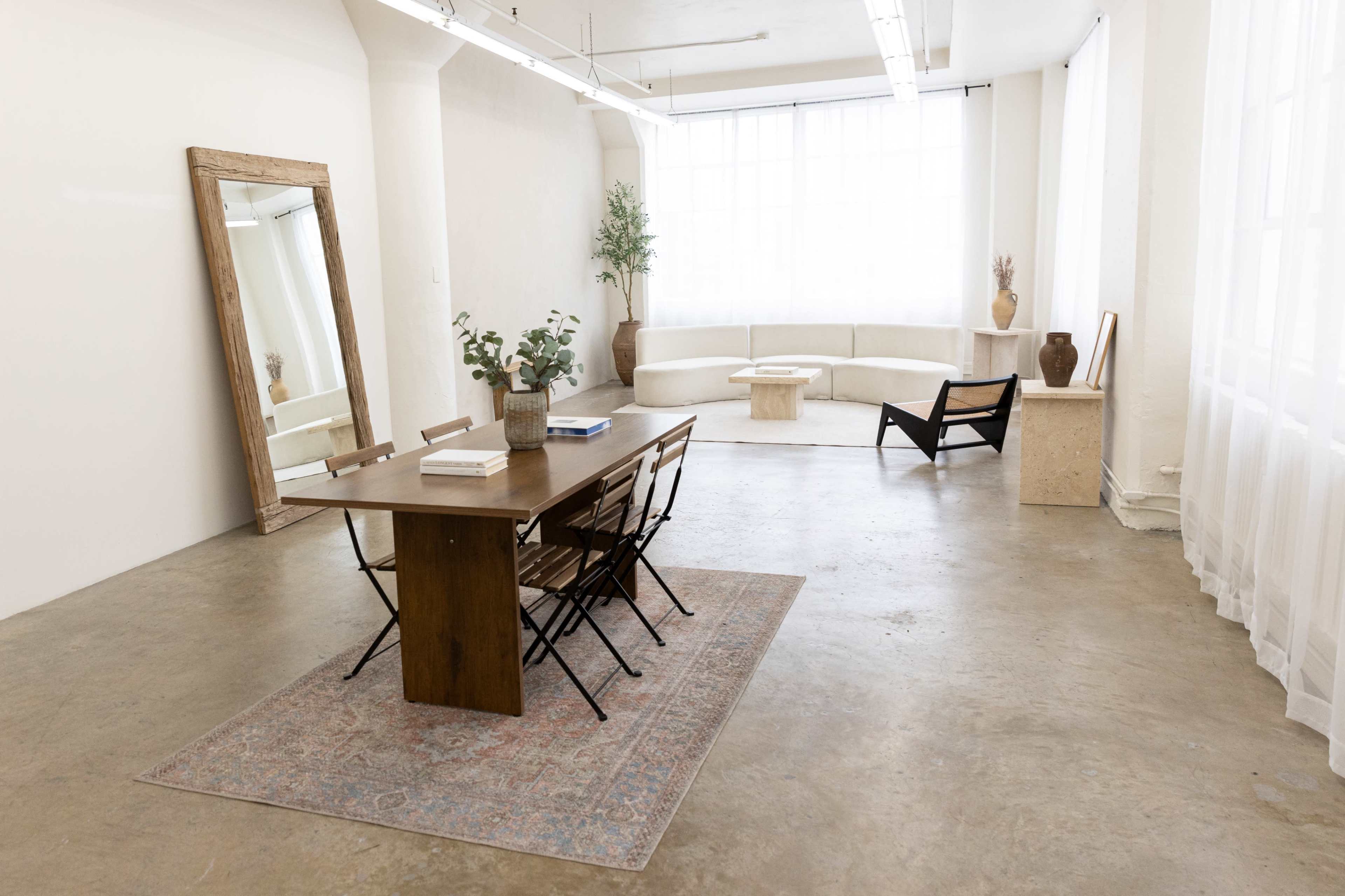 The image shows a minimalist room with a wooden dining table and chairs, large windows allowing natural light, and a modern seating area with a sofa and decorative elements.