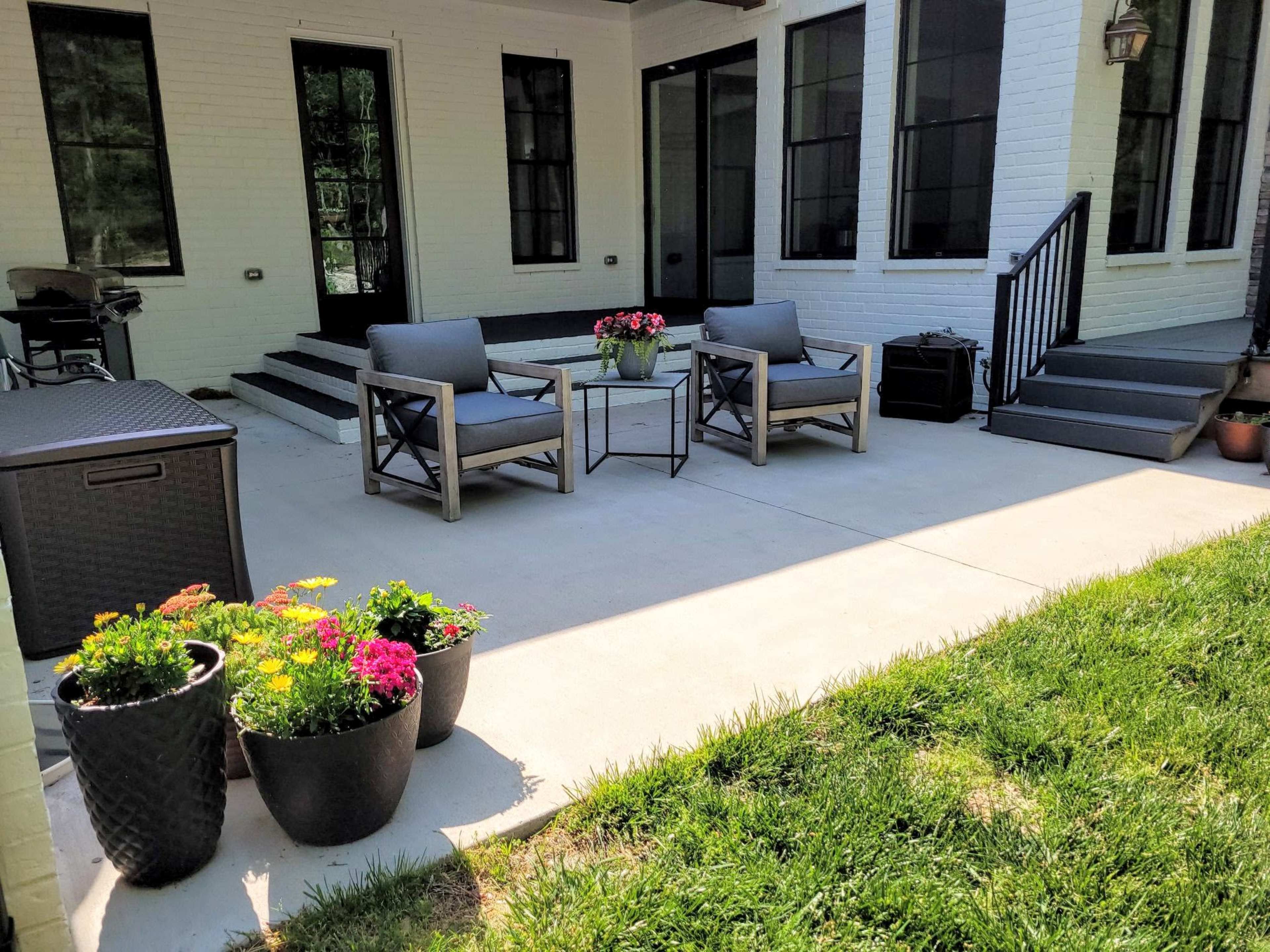 The image shows a patio area featuring two chairs and a small table, surrounded by potted plants and steps leading to a house with large windows.