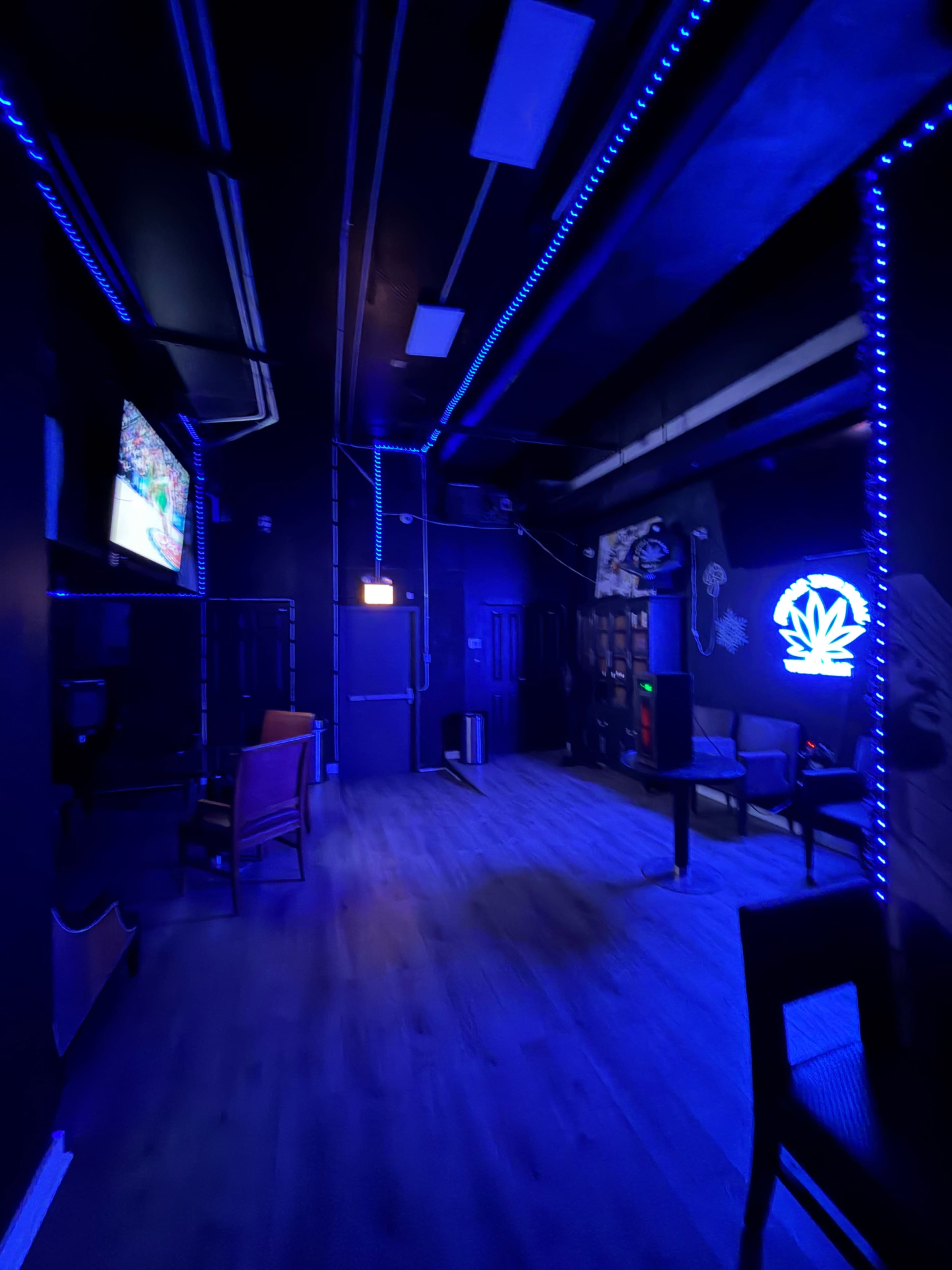 The image shows a dimly lit lounge area featuring blue LED lighting, a television screen, and sparse furniture including chairs and tables.