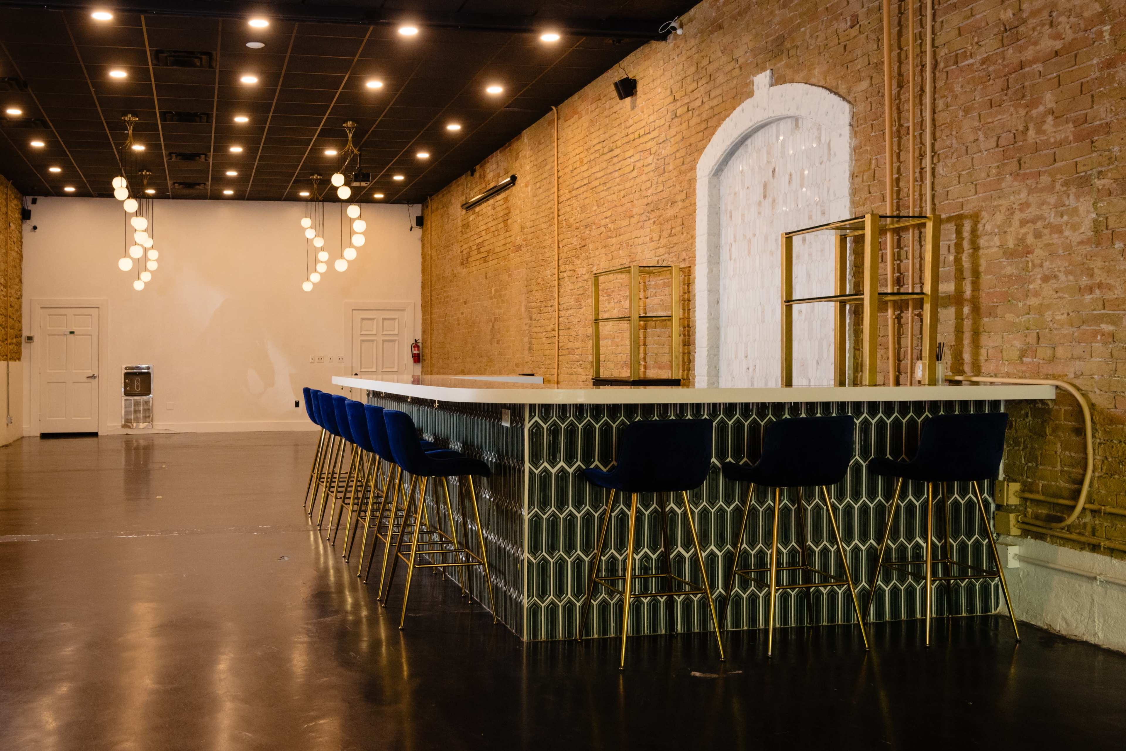 The image shows a spacious interior with exposed brick walls, featuring a bar with a patterned front and several blue bar stools arranged around it.